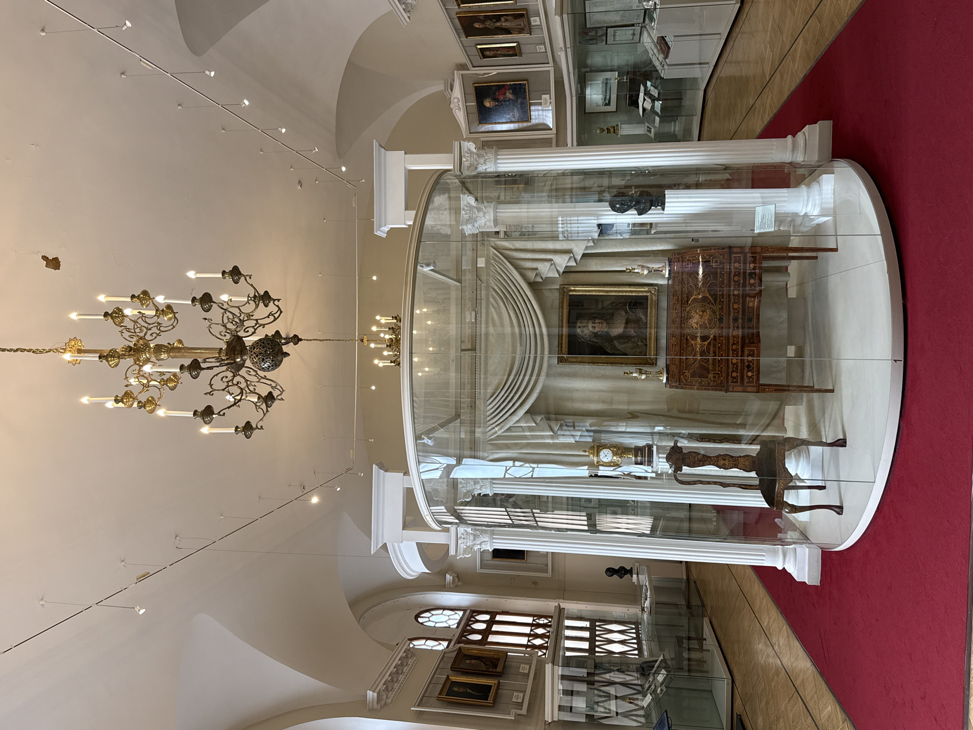 The image depicts the interior of a museum, likely in Moscow, Russia, featuring a central display encased in glass. This display contains an ornate antique desk adorned with intricate patterns and a framed portrait of a woman. To the left of the desk, a single antique wooden chair with carved details is positioned. Behind the desk and chair, beige drapes are artfully arranged, adding to the historical ambiance.
Flanking the glass enclosure are two classical white columns, supporting white architectural elements above. A grand, multi-tiered chandelier with candle-like lights hangs from the ceiling, illuminating the scene. Other exhibits are visible in the background, including framed paintings on the walls and glass display cases containing various artifacts. The floor is covered with a wooden parquet, with a vibrant red carpet leading towards the central display. The museum environment appears well-lit and meticulously maintained, showcasing historical artifacts within a classical architectural setting.