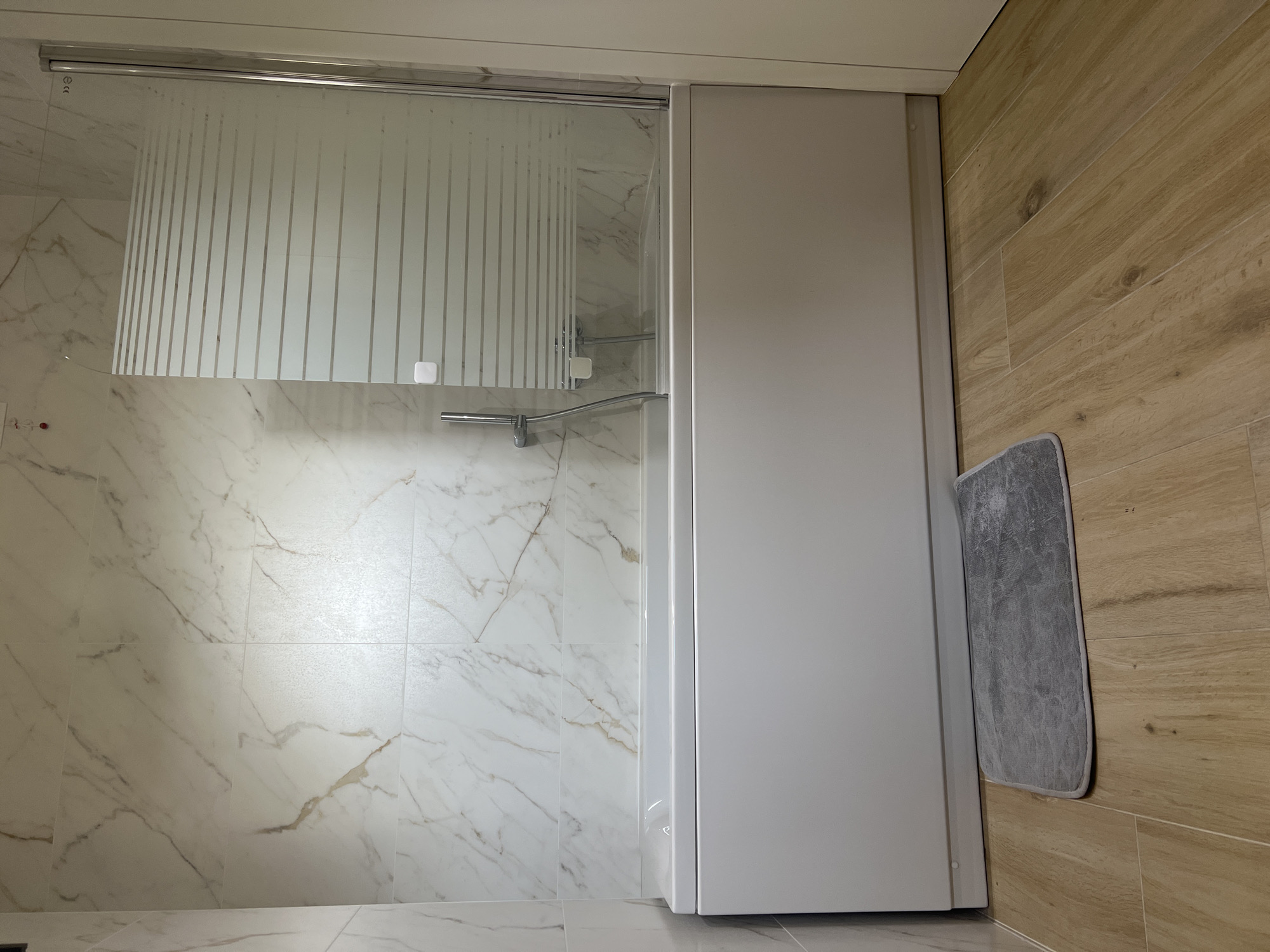 The image shows a bathroom with a white bathtub, a frosted glass shower screen, and marble-patterned tiles on the wall. A grey bath mat is placed on the wooden floor in front of the tub. A showerhead and faucet are visible on the wall behind the shower screen. The scene is static and there are no people present. This image is not related to Verona, Italy.