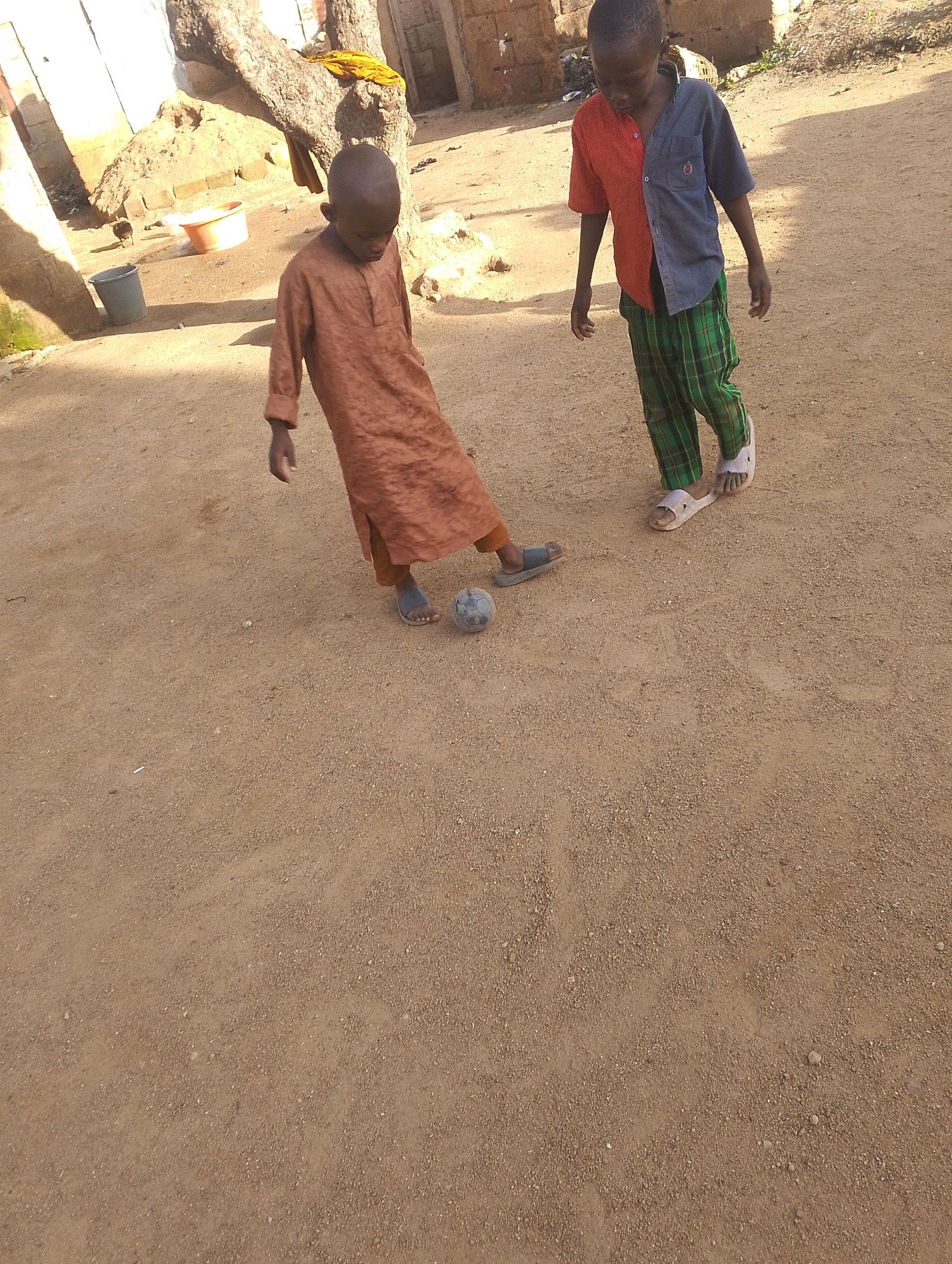 In a dirt yard in Unknown, Unknown, two young boys are playing with a soccer ball. One boy, wearing a brown tunic and sandals, is about to kick the ball. He is looking down at the ball, and his right foot is positioned to strike it. The other boy, wearing a two-toned shirt (half red, half blue) and green plaid pants with sandals, stands nearby, watching. The yard is dusty and dry, with a large tree trunk on the left side of the frame and some buildings in the background. There is a gray bucket and an orange basin near the tree. The lighting suggests it is daytime, possibly late afternoon due to the shadows.