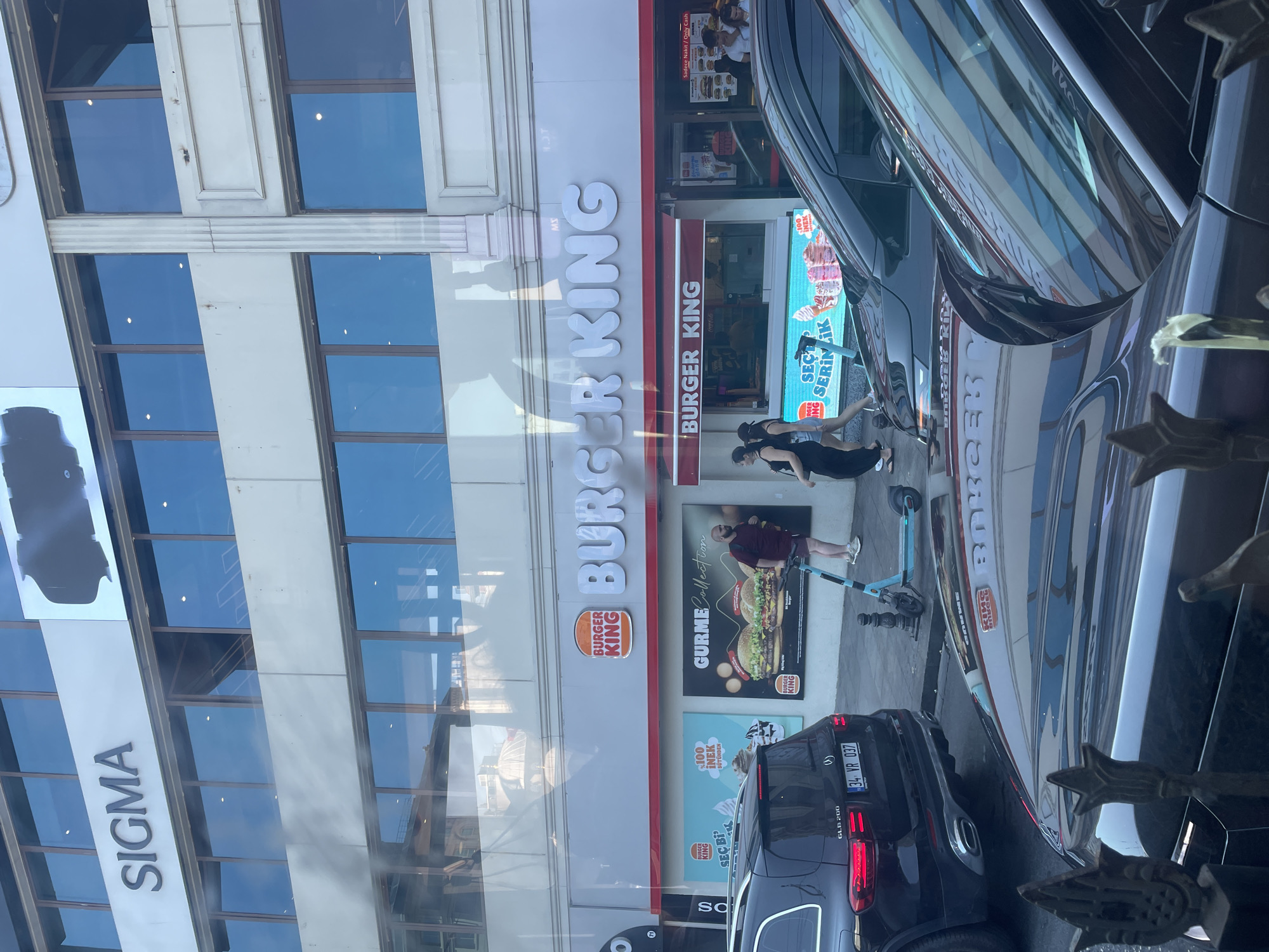 The image depicts a street scene in Istanbul, Turkey, with a prominent Burger King restaurant. The Burger King sign is clearly visible, featuring the iconic crown logo and white lettering against a light gray background. Below the main sign, a red awning stretches across the restaurant's facade. Advertisements are displayed on panels to the left and right of the Burger King entrance.
On the left side of the image, a black Mercedes-Benz GLB 200 is parked, its license plate reading "34 VR 037". To the right of the parked car, a turquoise electric scooter is standing on the sidewalk. Two women are walking past the scooter and the Burger King entrance. One is wearing a black sleeveless dress, and the other is in a black top and denim shorts.
The building above the Burger King features a large sign that reads "SIGMA" in bold, uppercase letters, also illuminated. The upper floors of the building have large windows.
Reflections are visible on the surfaces of the parked cars in the foreground, showing the Burger King sign and other elements of the streetscape, including traffic lights and the building opposite. The overall environment suggests a commercial area in a bustling city.