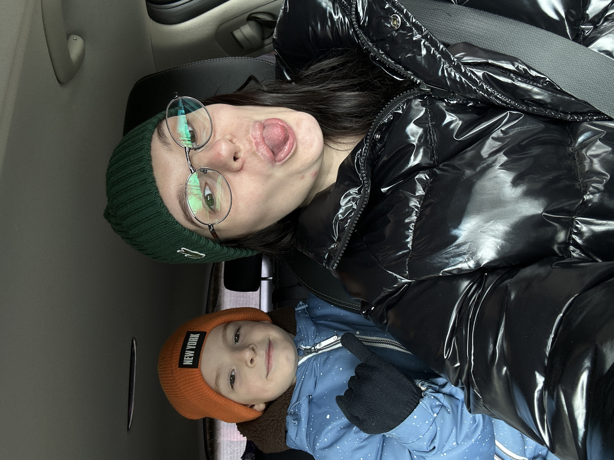 A woman with dark hair wearing a green beanie, round glasses, and a black puffer jacket is sticking her tongue out. A young boy wearing a brown beanie with the words "New York" on it and a blue jacket is smiling. They appear to be in a car with seatbelts on. The location is Прикамский, Russia.
