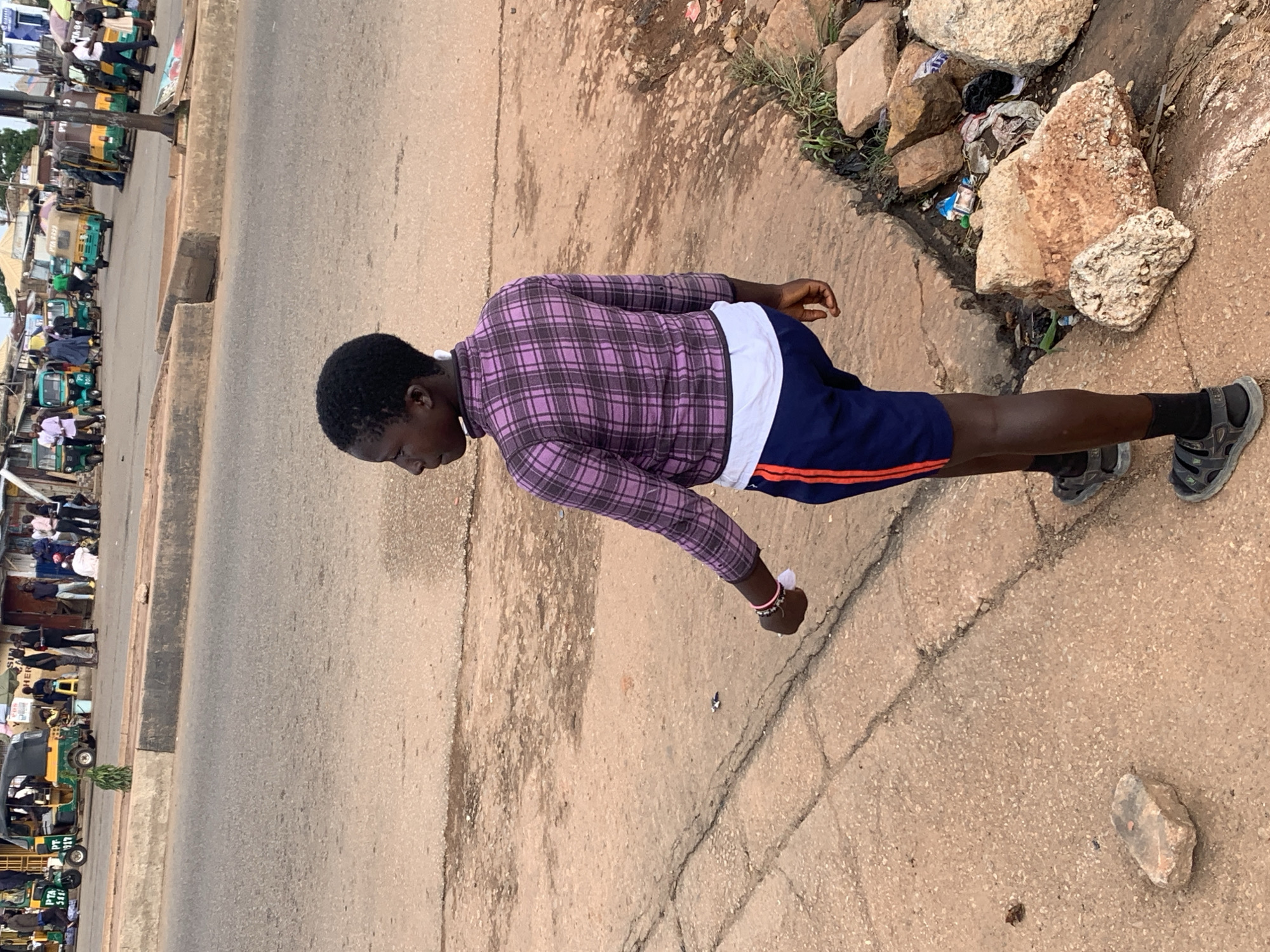 The image depicts a street scene in Gura Top, Nigeria. In the foreground, a young person wearing a purple plaid long-sleeved shirt over a white t-shirt, blue shorts with a red stripe, black socks, and sandals, walks away from the camera along a paved walkway. A small white piece of paper is visible in their right hand. The background features a dusty road with several auto-rickshaws, commonly known as "keke napep," parked and in motion. Numerous people are scattered around the area, some standing, some sitting, and some appearing to be involved in commercial activities near the vehicles. The environment appears to be a bustling market or transport hub, with a rough, unpaved section of the street on the right side of the image containing rocks and scattered refuse. The overall impression is one of a busy, everyday street scene in a Nigerian city.