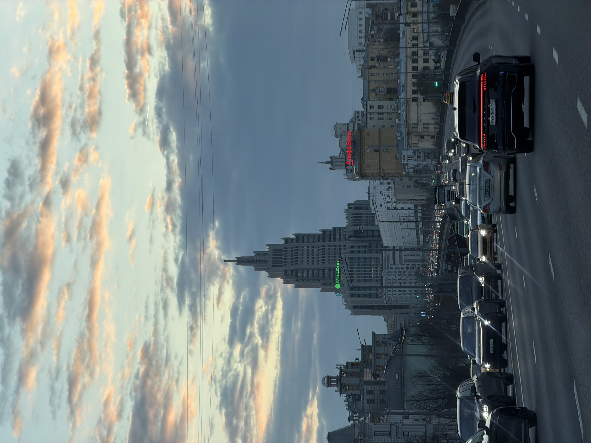 The image captures a traffic jam in Moscow, Russia, juxtaposed against the backdrop of a pastel sky at sunset. In the foreground, numerous cars are stuck on a multi-lane highway. Prominently, the rear of a black car with illuminated taillights is in the right side of the shot. Behind the traffic, the urban landscape features a mix of older buildings and modern high-rises, including a tall skyscraper with a distinctive spire, recognizable as one of the Seven Sisters. Signage for "Альфа-Банк" (Alfa-Bank) and "Мегафон" (MegaFon) are visible on buildings, identifying the location as Moscow. The sky is filled with elongated, golden-edged clouds set against a backdrop of blue and grey. Power lines run across the sky.
