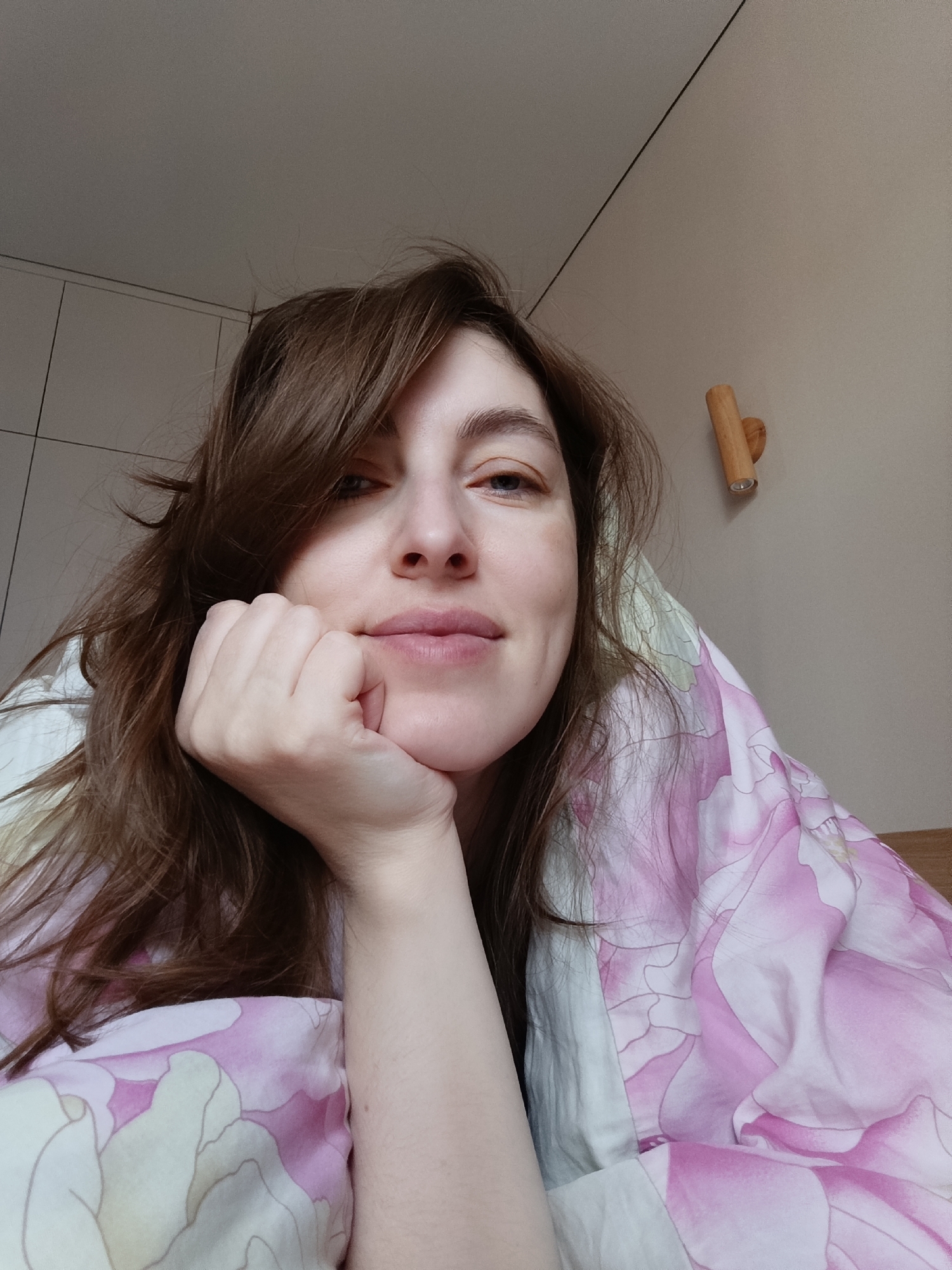 A woman with brown hair is lying in bed, propped up on her left hand, smiling slightly at the camera. She is under a pink and white floral comforter. A wooden sconce is mounted on the pale wall behind her to the right. In the background, there is a closet with white doors. The location of the photo is not discernible from the image. There is no indication it was taken in the city of Chaikovsky, Russia.