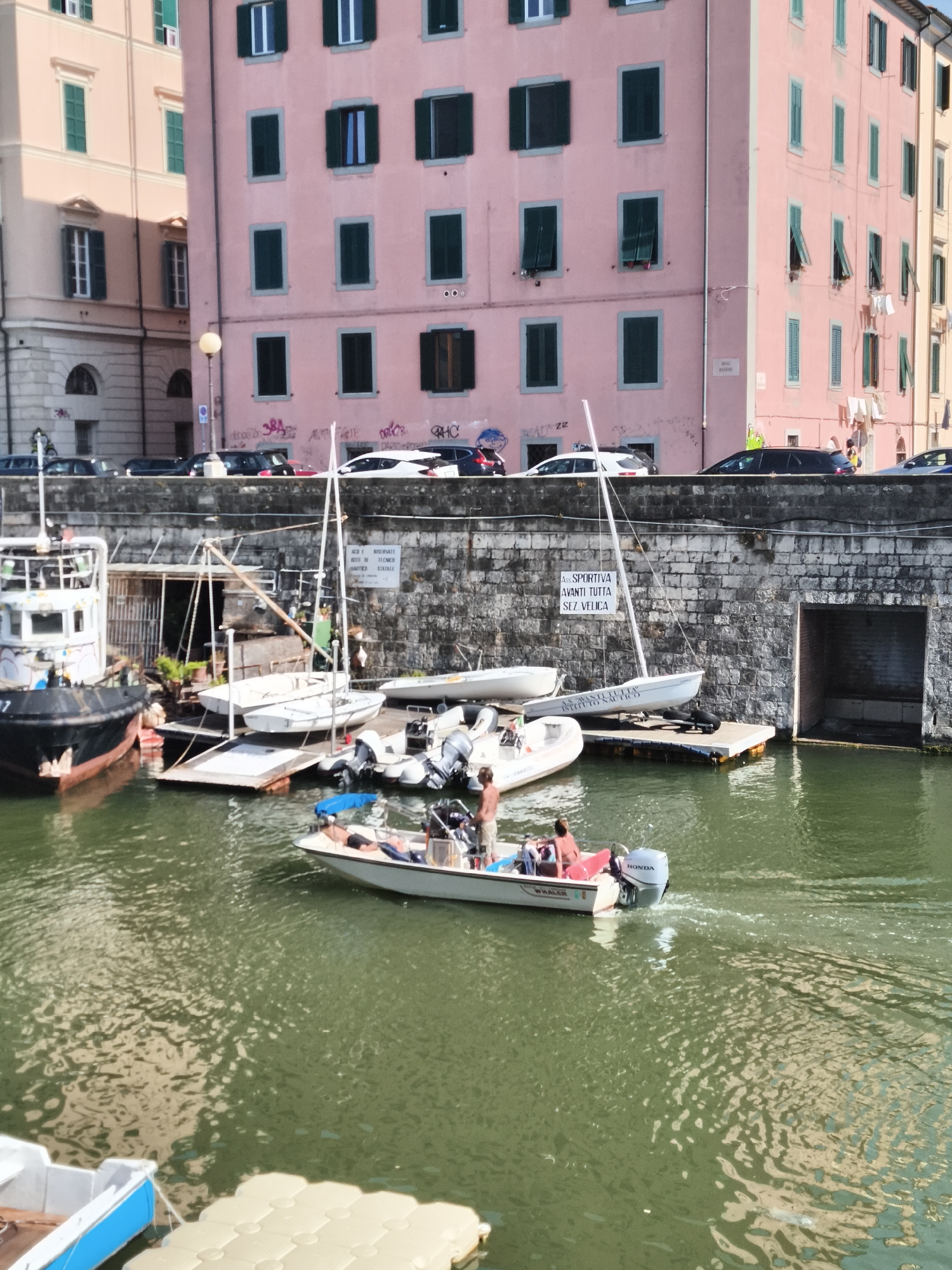 This image captures a sunlit canal scene in Livorno, Italy, likely near the historic Darsena area given the distinctive pink buildings and the presence of docked boats. A multi-story pink building with numerous closed green shutters dominates the background, its facade showing signs of age and some graffiti. Adjacent to it is another building, lighter in color, also featuring green shutters.
In the foreground, the canal's murky green water reflects the sunlight and the buildings. Several boats are visible. A larger tugboat, painted black and white with graffiti, is docked on the left. Beside it, smaller white sailboats and motorboats are moored. One motorboat, with a visible "HONDA" engine, is actively moving, creating ripples in the water. There are at least three individuals visible in this motorboat: one standing, and two seated. One of the seated individuals appears to be lying down. Another smaller boat with a blue trim is partially visible in the lower left corner, suggesting it is also moored.
A stone wall runs along the canal's edge, with an arched opening on the right side, possibly leading to an indoor area. A white sign with black text is affixed to the stone wall, partially obscured by a sailboat's mast. The overall atmosphere is one of a working harbor or marina, with a mix of leisure and possibly commercial activity. The presence of laundry hanging from balconies on the pink building suggests residential use, adding a touch of everyday life to the scene.