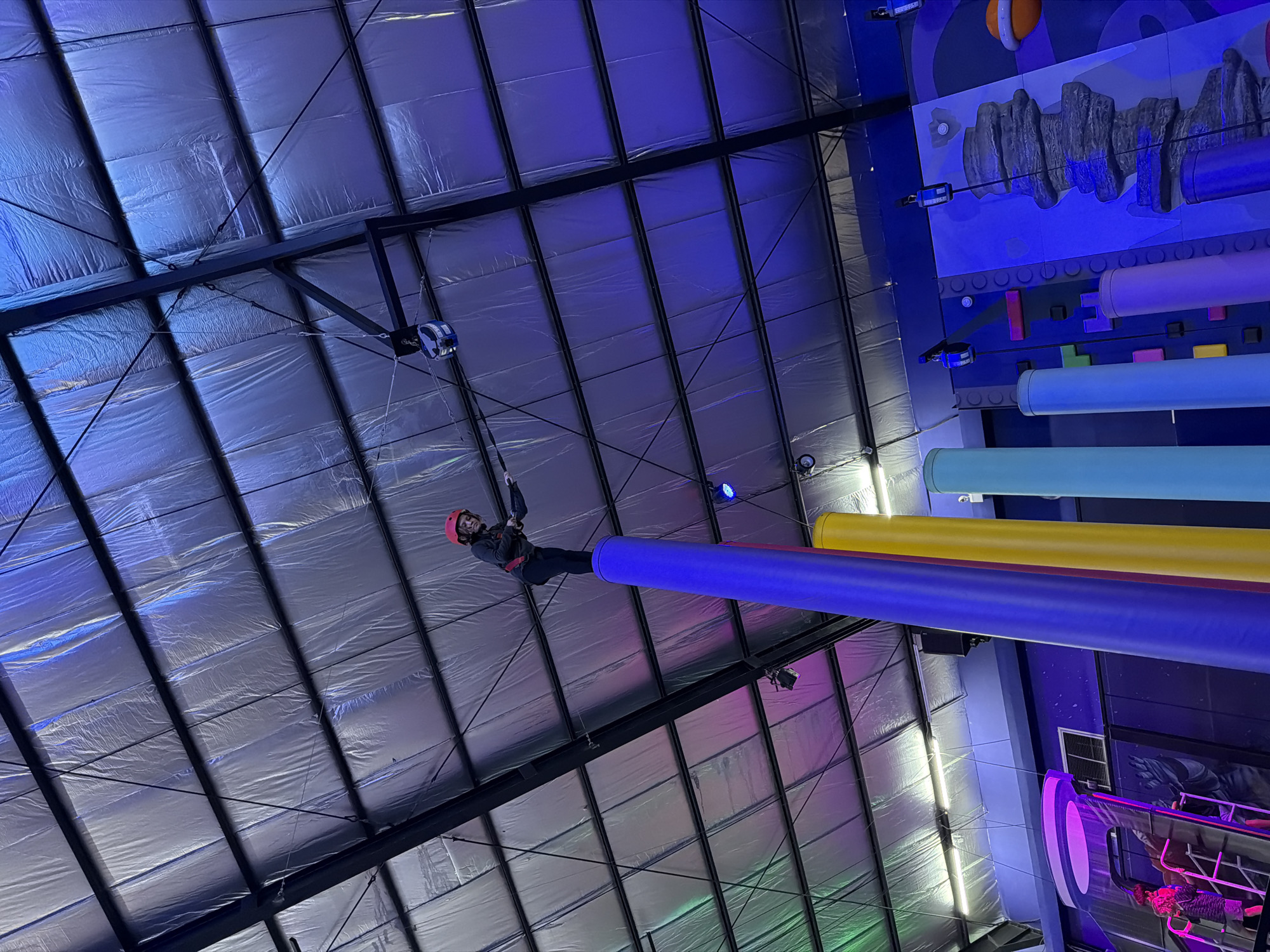A low-angle shot captures a person in mid-air, suspended by cables, in an indoor adventure park. The individual, wearing a dark jacket and a red helmet, is positioned horizontally, with their body almost parallel to a series of large, colorful cylindrical obstacles. These obstacles are aligned vertically, in hues of blue, yellow, and light blue. The park's interior has a high ceiling with a grid-like metal structure, covered with reflective silver panels. In the background, a blue-toned wall displays decorative elements, including a textured rock-like formation and circular shapes. The scene is illuminated by various light sources, casting shadows and creating a dynamic play of light and shadow on the ceiling and the person in motion. The location is Denpasar, Indonesia.