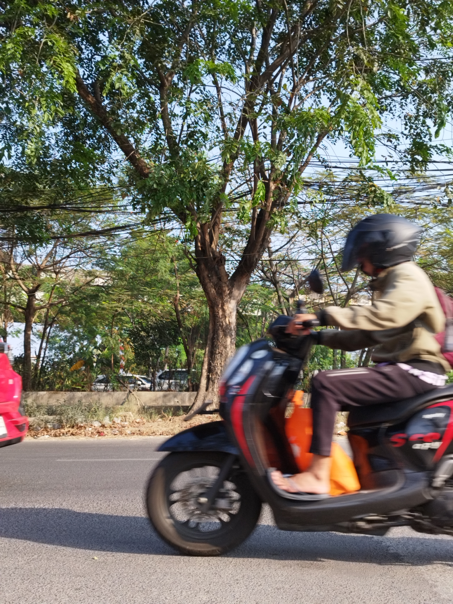 An eye-level, medium shot shows a person riding a scooter on a paved road in an outdoor setting. The scooter, which is black with red accents, is in motion, creating a blur effect. The rider, wearing a black helmet and a light brown long-sleeved shirt, is seated on the scooter. They are also wearing dark pants and appear to be barefoot. An orange object is visible in the footwell of the scooter. To the left of the scooter, the rear of a red car is partially visible. In the background, a large tree with green foliage stands prominently, with its branches extending upwards. Power lines are strung between poles behind the tree. Further in the background, there is more greenery, suggesting a park or a roadside with trees. The sky is blue and appears clear. The overall scene depicts a typical urban environment with active transportation.
