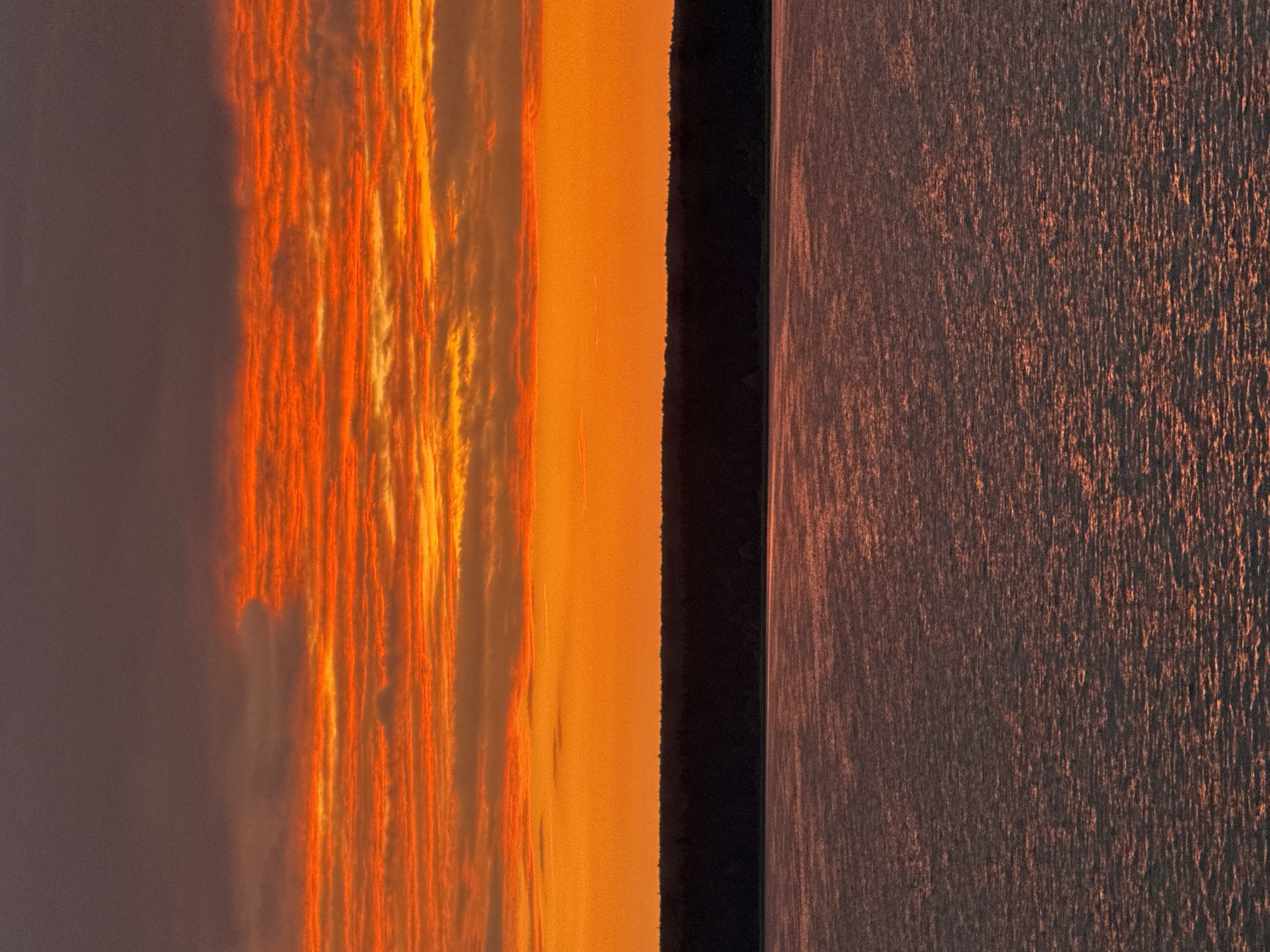 The image depicts a sunset over a body of water, likely a reservoir or a wide river, bordered by a dark, dense line of trees on the horizon. The sky is dominated by vibrant hues of orange and red, with layered clouds creating a textured effect. The upper portion of the sky transitions into a darker, purplish-grey, suggesting the last moments of daylight. The water in the foreground reflects the colors of the sky, creating shimmering patterns of light and shadow on its surface. The scene is devoid of any human presence, structures, or identifiable landmarks within the city of Votkinsk, Russia. The overall atmosphere is one of natural beauty and tranquility during twilight.