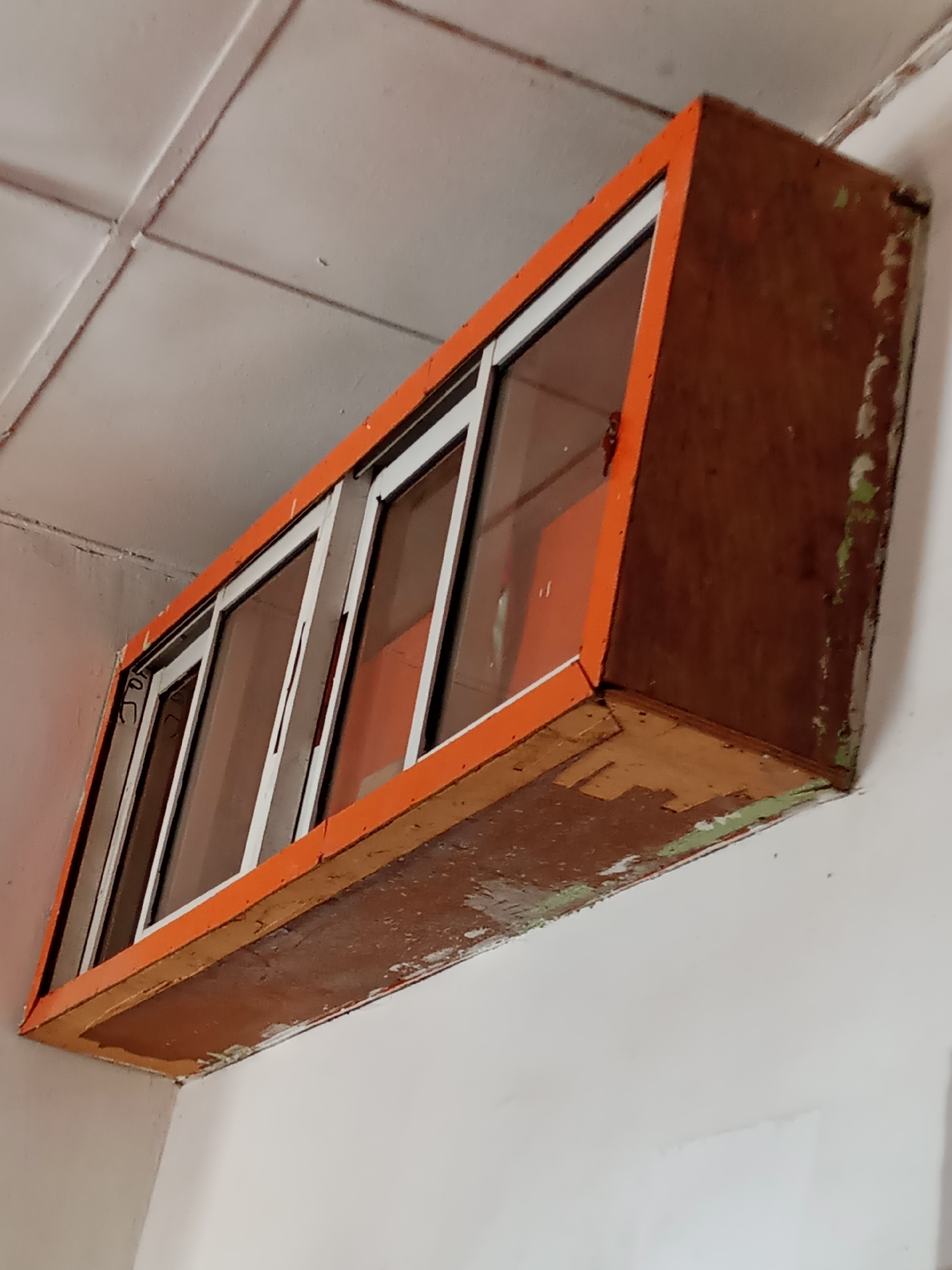 An orange-trimmed cabinet with sliding glass doors is mounted on a white ceiling. The cabinet appears to be made of wood and shows signs of wear, with peeling paint on its underside. The interior of the cabinet is also orange. The scene is indoors, likely in a room with white walls. There are no people visible in the image, and no specific activity is taking place. The lighting suggests daytime, but there are no other cues about the time of day, weather, or specific location beyond Jalingo, Nigeria. Some faint markings are visible on the glass doors, but they are too indistinct to be read.