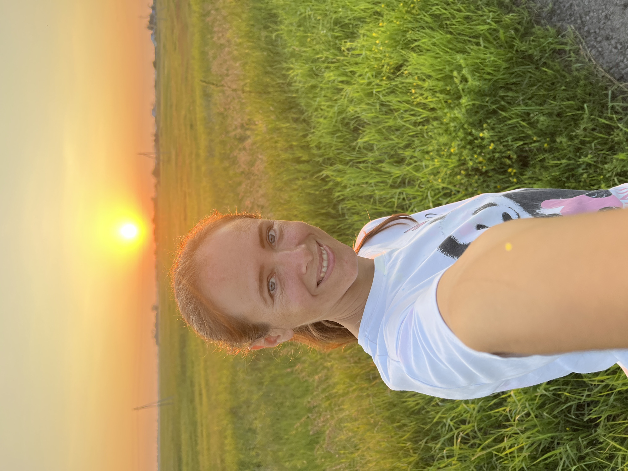 A woman with brown hair and blue eyes is taking a selfie in a grassy field at sunset. She is wearing a white t-shirt with a panda graphic. The sun is low on the horizon, casting a warm orange glow over the scene. In the background, there are utility poles and what appear to be distant buildings. The overall environment is rural, with tall green grass and a clear sky. The image was taken in Pivkino, Russia.