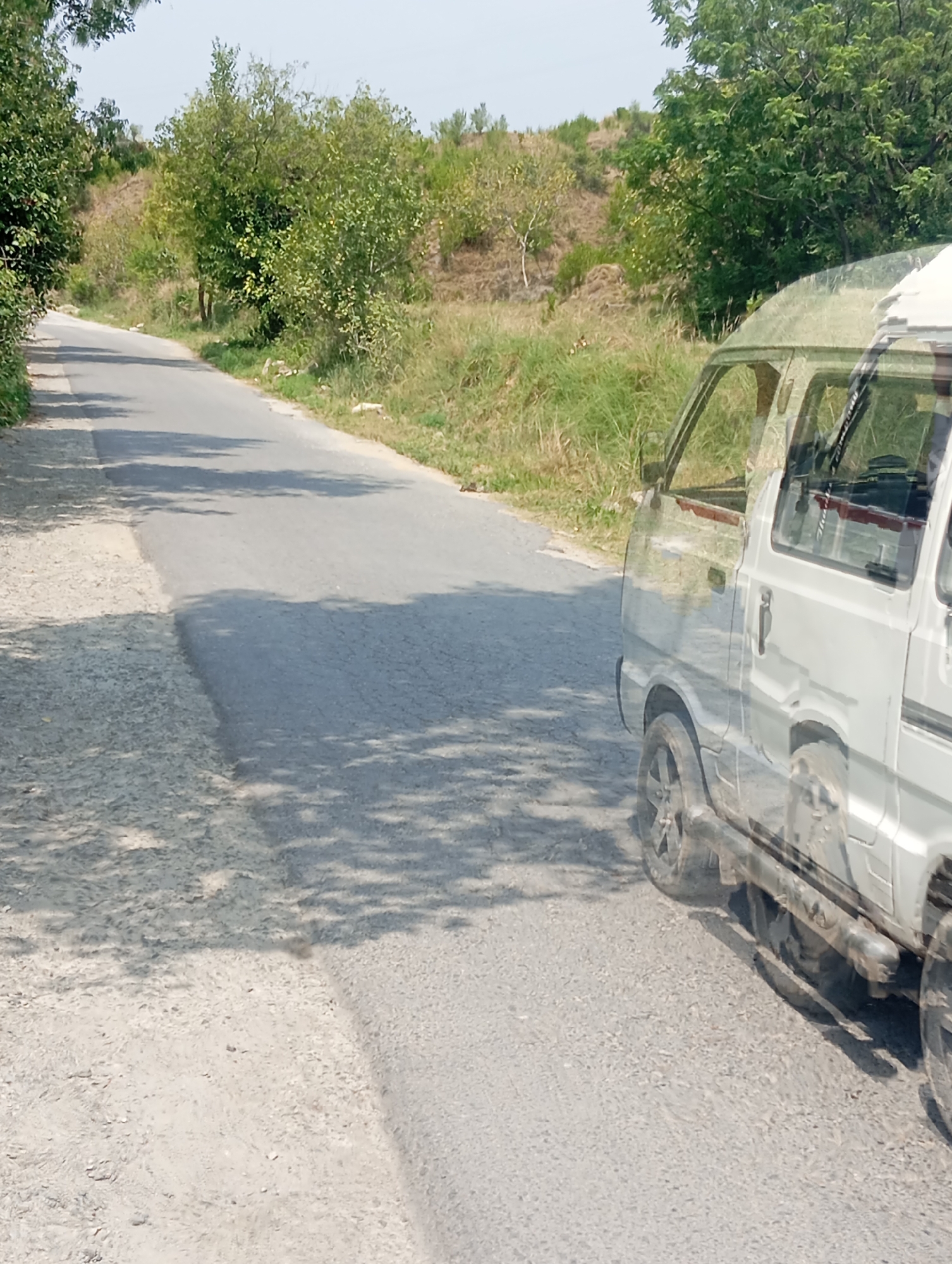 The image shows a two-lane road with a rough, light-colored shoulder on the left. The road surface is asphalt and shows signs of wear. Lush green trees and vegetation line the right side of the road, with a hilly terrain in the background. The sky is clear and bright, suggesting a sunny day. A white van, partially transparent and layered with reflections of the surrounding environment, is visible on the right side of the road, appearing to be in motion. The van's wheels and lower body are distinctly visible, along with parts of its windows and roof. The overall scene depicts a rural or semi-rural road in Lassan, Pakistan, during daylight hours.