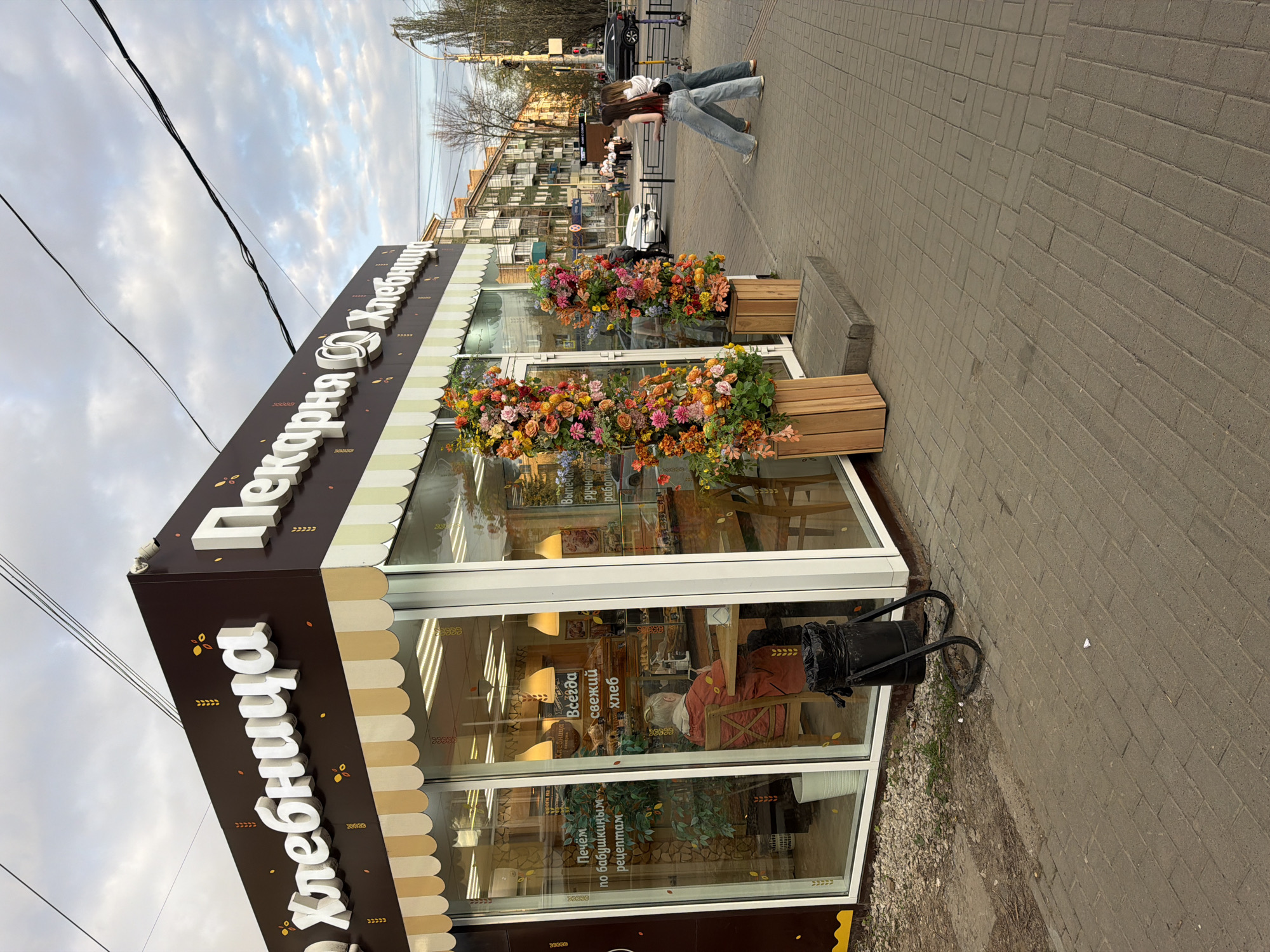 The image shows the exterior of a bakery named "Khlebnitsa" in Izhevsk, Russia. The bakery, identifiable by its brown and white signage with the name in Russian, has large windows displaying its interior, where seating and decorative lamps are visible. Two large flower boxes with brightly colored flowers flank the entrance. A black trash can is positioned near the bakery's side. The scene is set on a brick sidewalk, and two women are walking nearby in the background along with other pedestrians and vehicles visible down the street, indicating an urban setting. The sky is overcast.