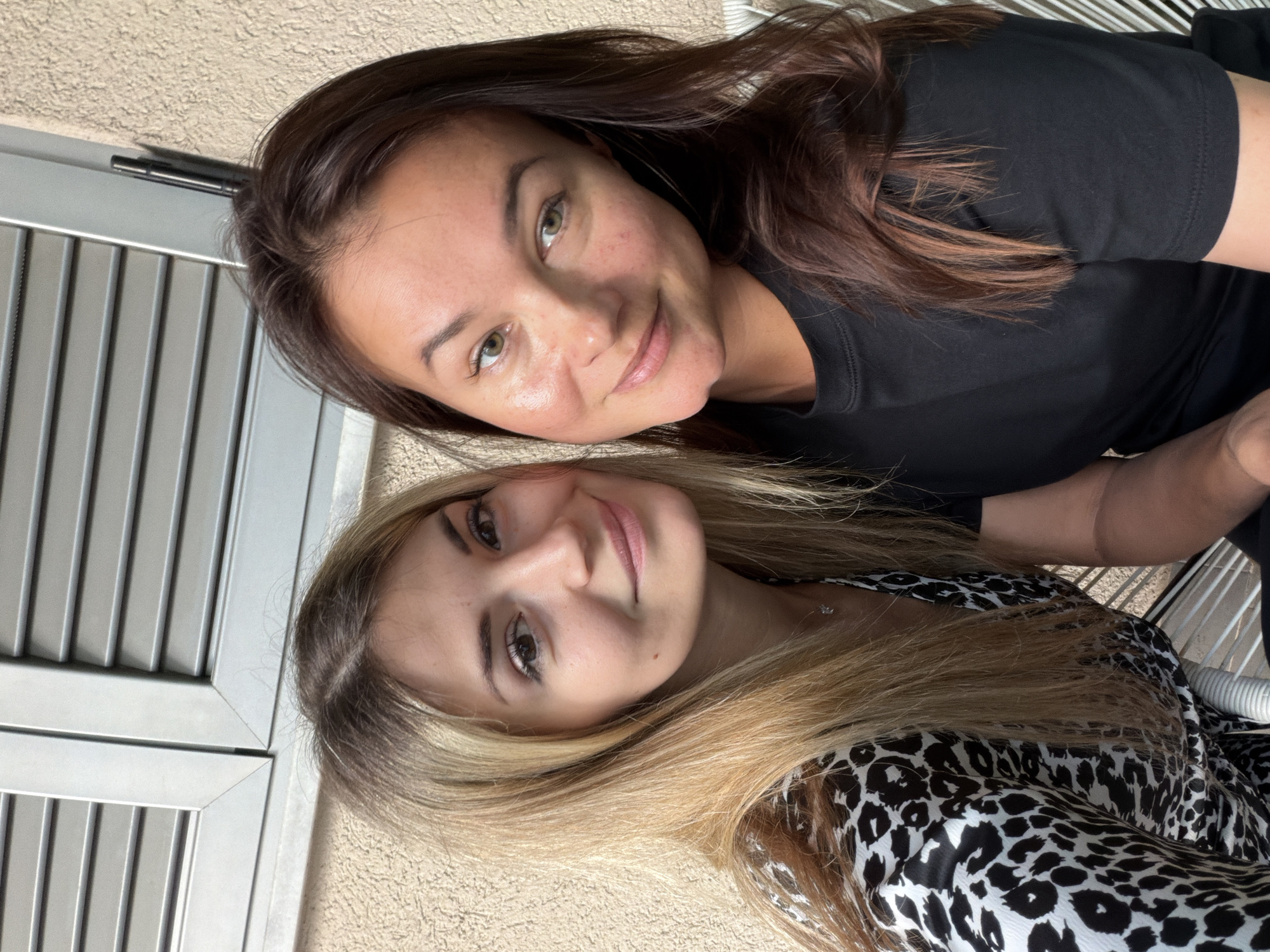 Two women are posing for a selfie in Budva, Montenegro. The woman on the top has brown hair, light skin, and is wearing a black shirt. The woman on the bottom has blonde hair, light skin, and is wearing a black and white animal print dress. They are sitting outside, next to a shuttered gray window. The exterior walls of the building are beige.