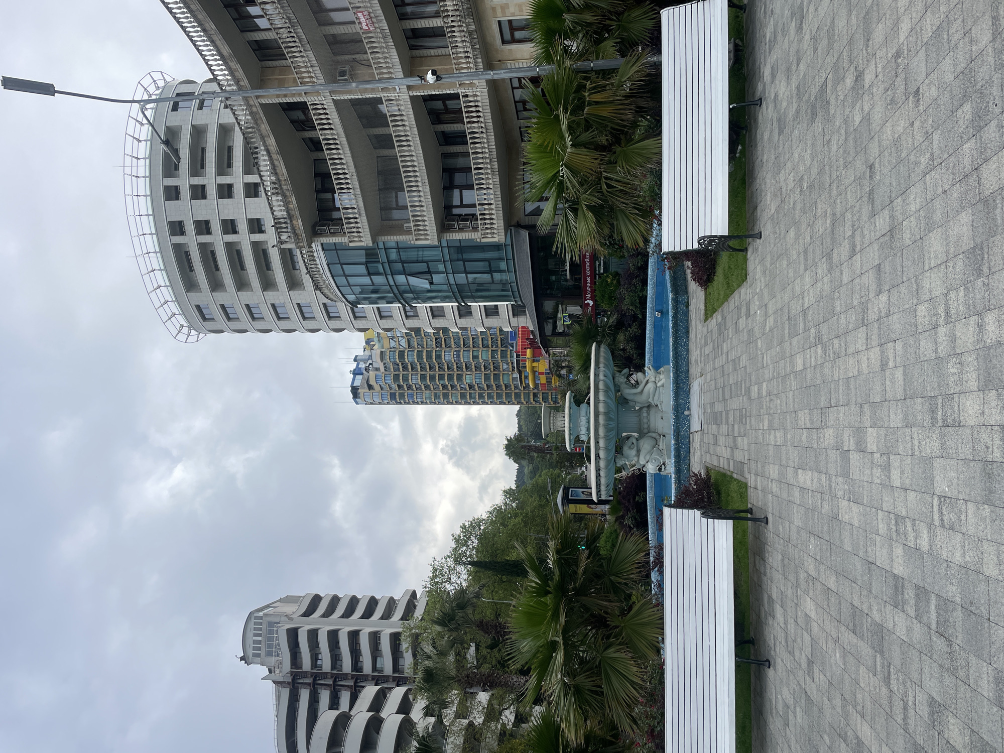 The image depicts a cityscape, likely a section of Sochi, Russia, with distinctive architecture. A paved pedestrian area occupies the right portion of the frame, lined with white benches. To the left of the walkway is a fountain. Buildings are interspersed with palm trees and other greenery. The sky is overcast. The buildings exhibit unique rounded and tiered designs. The architectural style suggests a modern, urban environment.