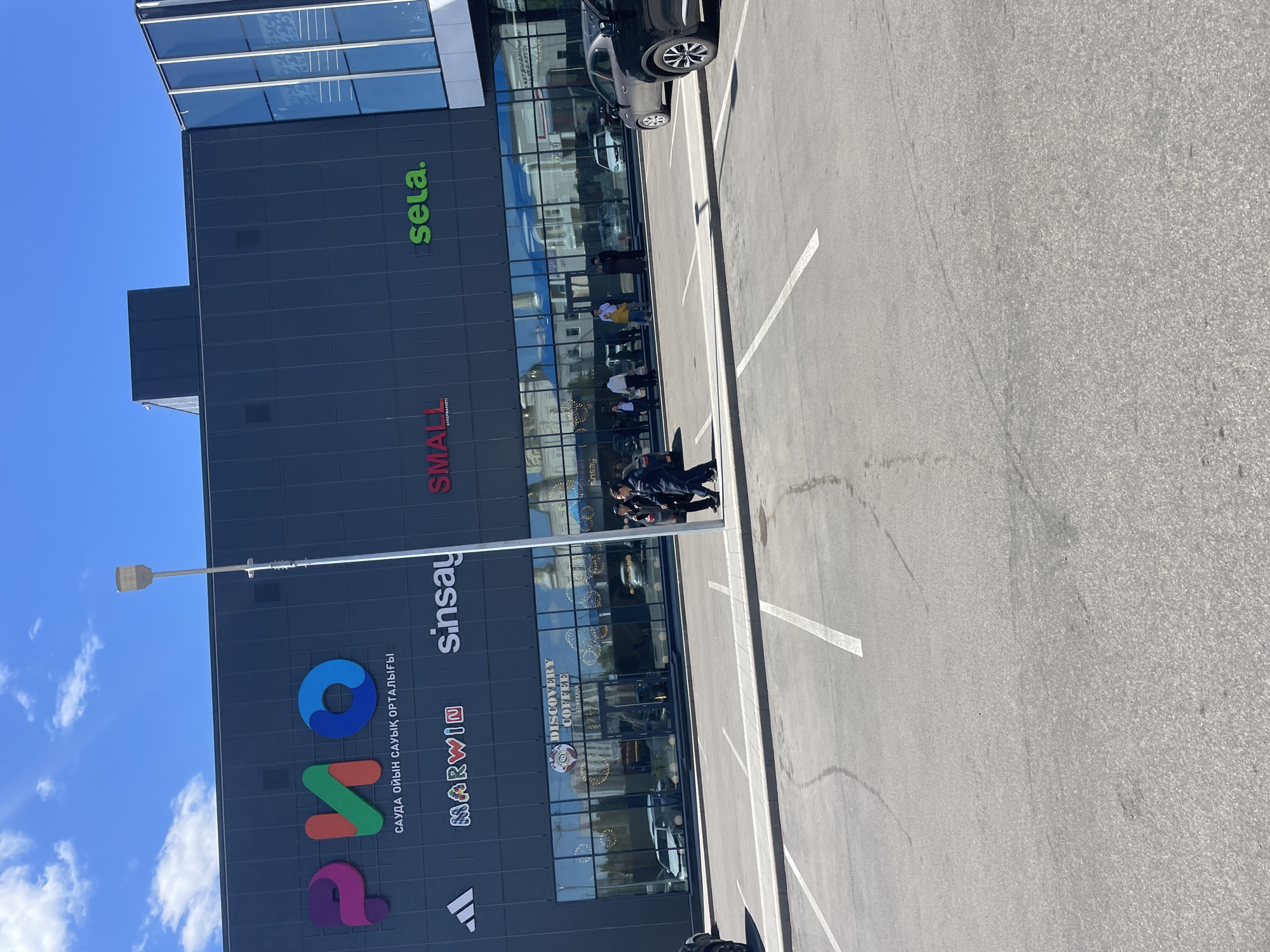 A low-angle daytime shot captures a building facade with large vertical signage in Kokshetau, Kazakhstan. The building features brands such as Adidas, Marwin, Sinsay, Small, and Sela. People are visible in front of the building, and a parking lot is visible on the right side. The sky above is blue with scattered clouds.