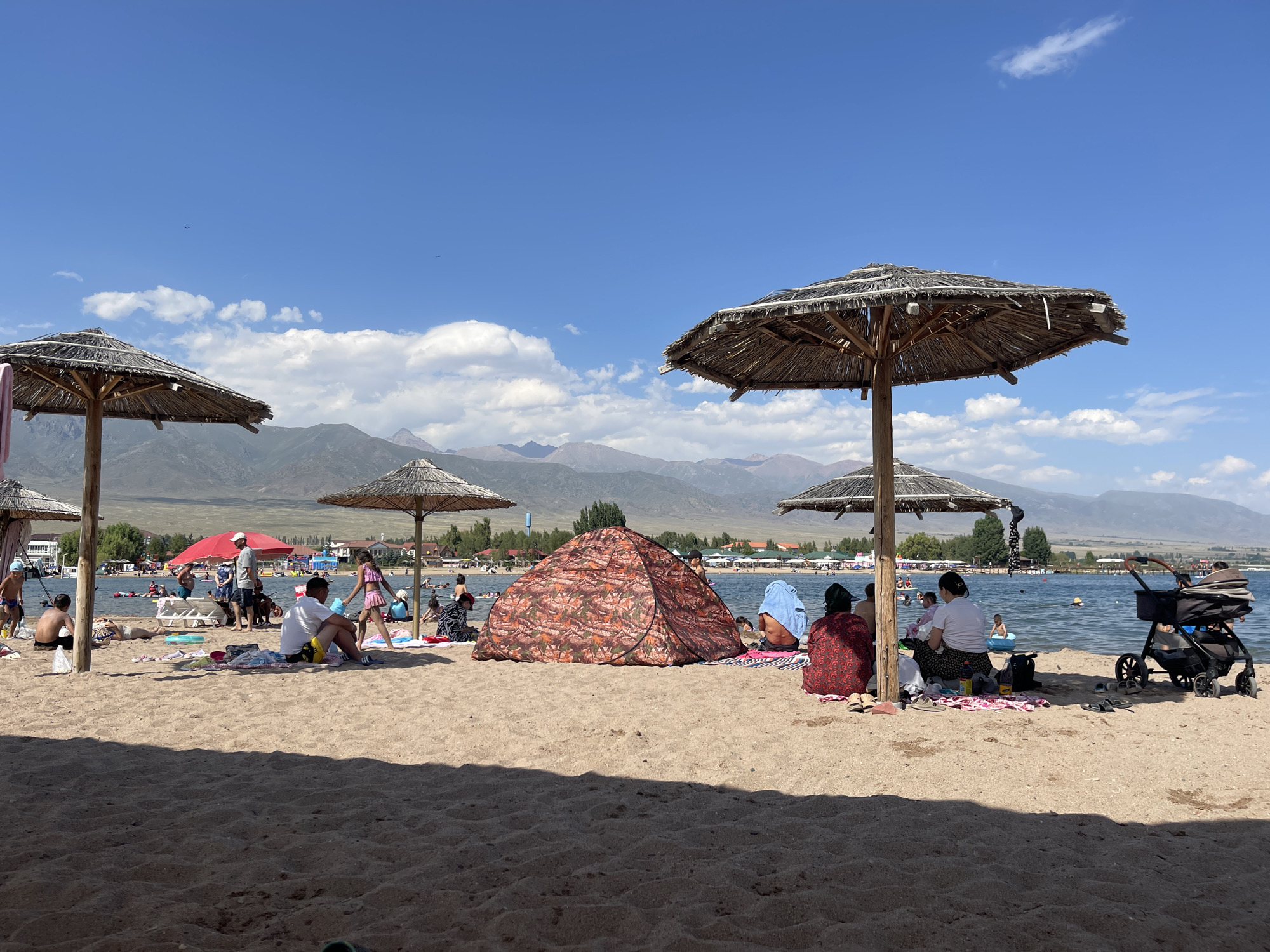 The image depicts a sunny day at a beach located in Choktal, Kyrgyzstan. The foreground is dominated by sand, with several people scattered across it. In the mid-ground, a lake stretches out, with numerous individuals swimming and enjoying the water. Beyond the lake, a line of trees and buildings marks the shoreline, leading to a backdrop of majestic mountains under a clear blue sky dotted with fluffy white clouds.
Several thatched umbrellas provide shade on the beach. Under one of these umbrellas, a group of people are gathered, some seated on blankets. In the center of the beach, a patterned tent is set up, and nearby, a family is picnicking. To the right, a baby stroller is parked on the sand.
The people are dressed in casual beach attire. Some are lounging, while others are interacting with each other or with children. The overall atmosphere is one of leisure and recreation on a warm summer day. The presence of mountains in the background and the style of buildings suggest a location in a region known for its natural beauty.