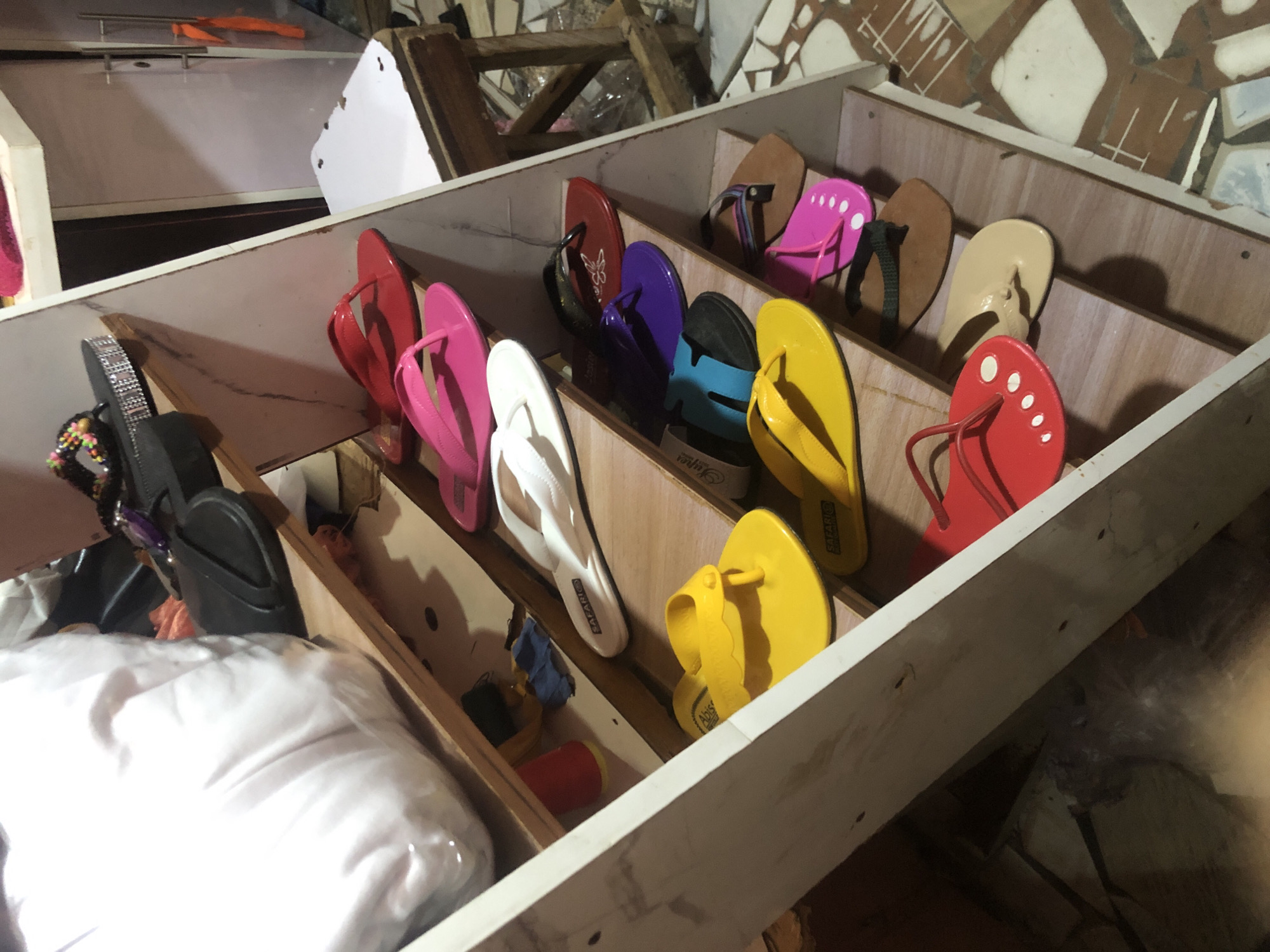 The image depicts a display of various flip-flops and sandals arranged on angled wooden shelves. The items are predominantly women's footwear, with a variety of colors and styles including red, pink, yellow, white, purple, blue, and brown. Some of the flip-flops have decorative elements, such as beads or embossed patterns. A large white object, possibly a bag or cushion, is visible on the top shelf. The background suggests an indoor retail environment, with elements of shelving and possibly a doorway or cabinet visible. The overall scene suggests a shoe store or market stall. The specific location in Ado, Nigeria, cannot be determined from the visual information provided in the image.