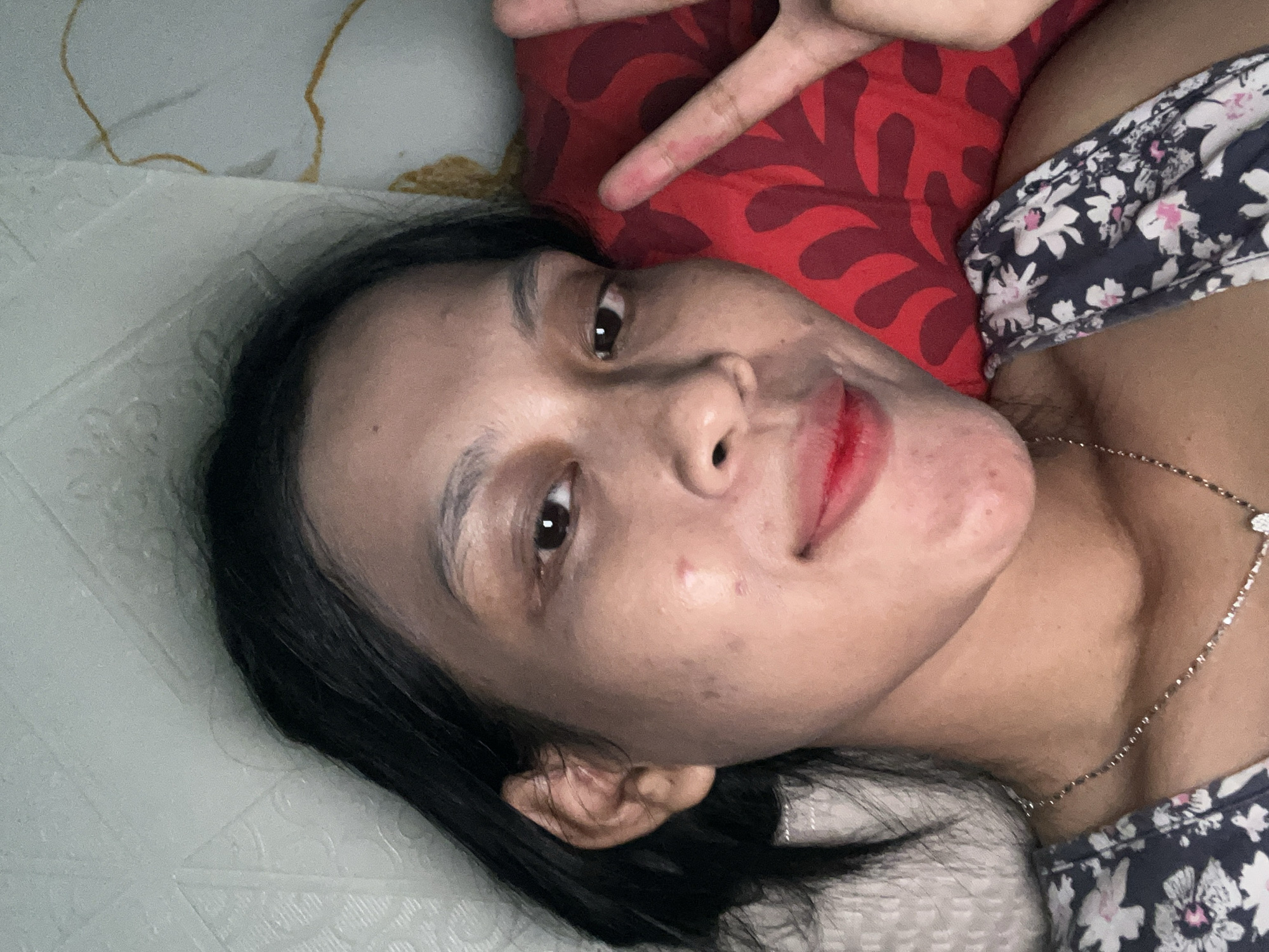 A young woman is lying down, looking at the camera. She has dark hair and is wearing a floral-patterned top and a silver necklace. Her left hand is raised, fingers splayed in a peace sign. She is wearing red lipstick. The background appears to be a bed with a red pillow. The location is identified as Kecamatan Pati, Indonesia.