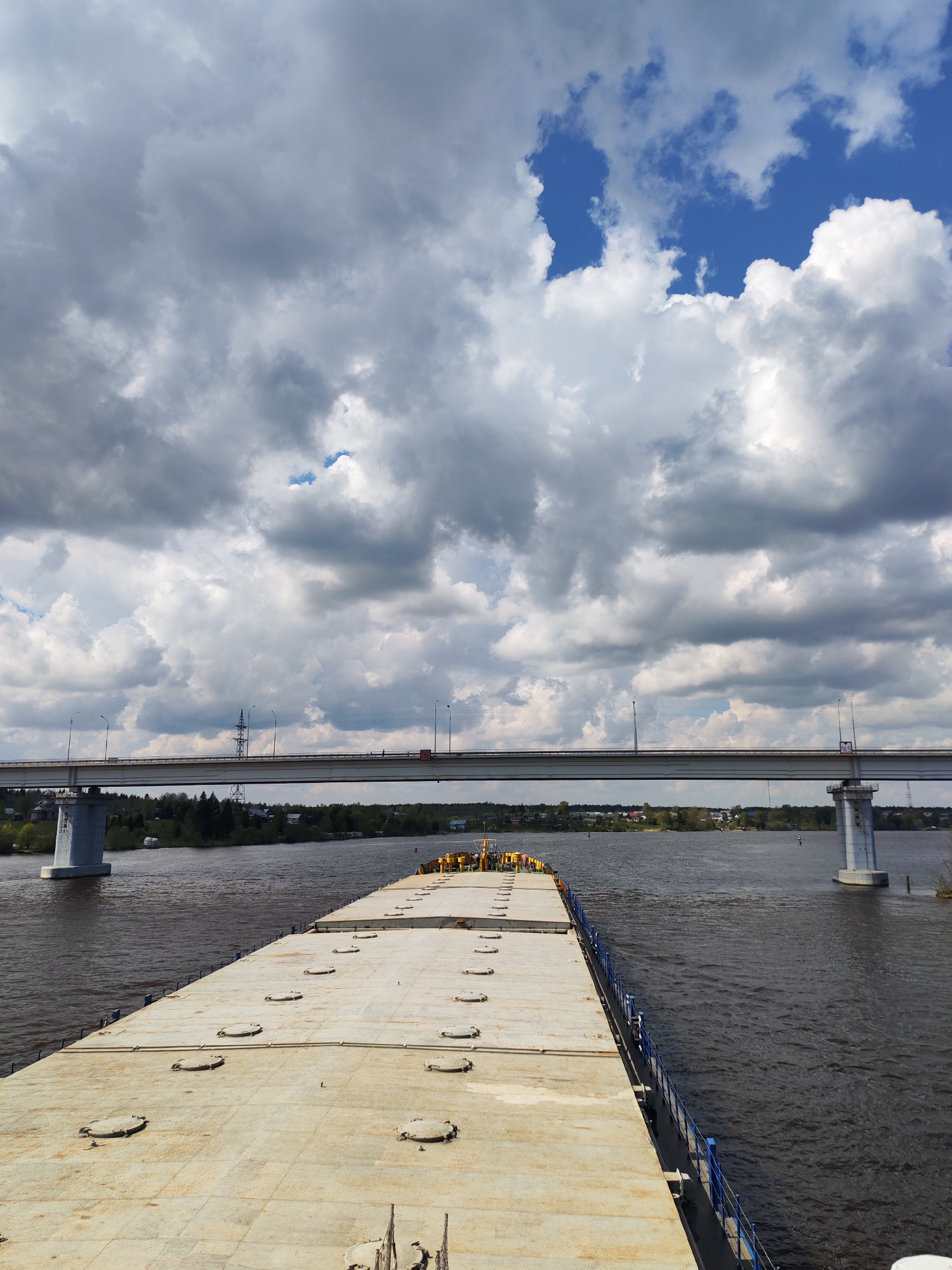 The image shows a barge sailing on a body of water with a bridge overhead. The barge has a flat, weathered surface with multiple circular hatches. A blue railing lines the edge of the barge. The water is dark and rippled. The bridge is supported by large concrete pillars and carries a road with streetlights and a power line structure. The shoreline is visible in the distance, featuring trees and some buildings. The sky is filled with a mix of dark, heavy clouds and patches of blue sky. The location is Unknown, Unknown.