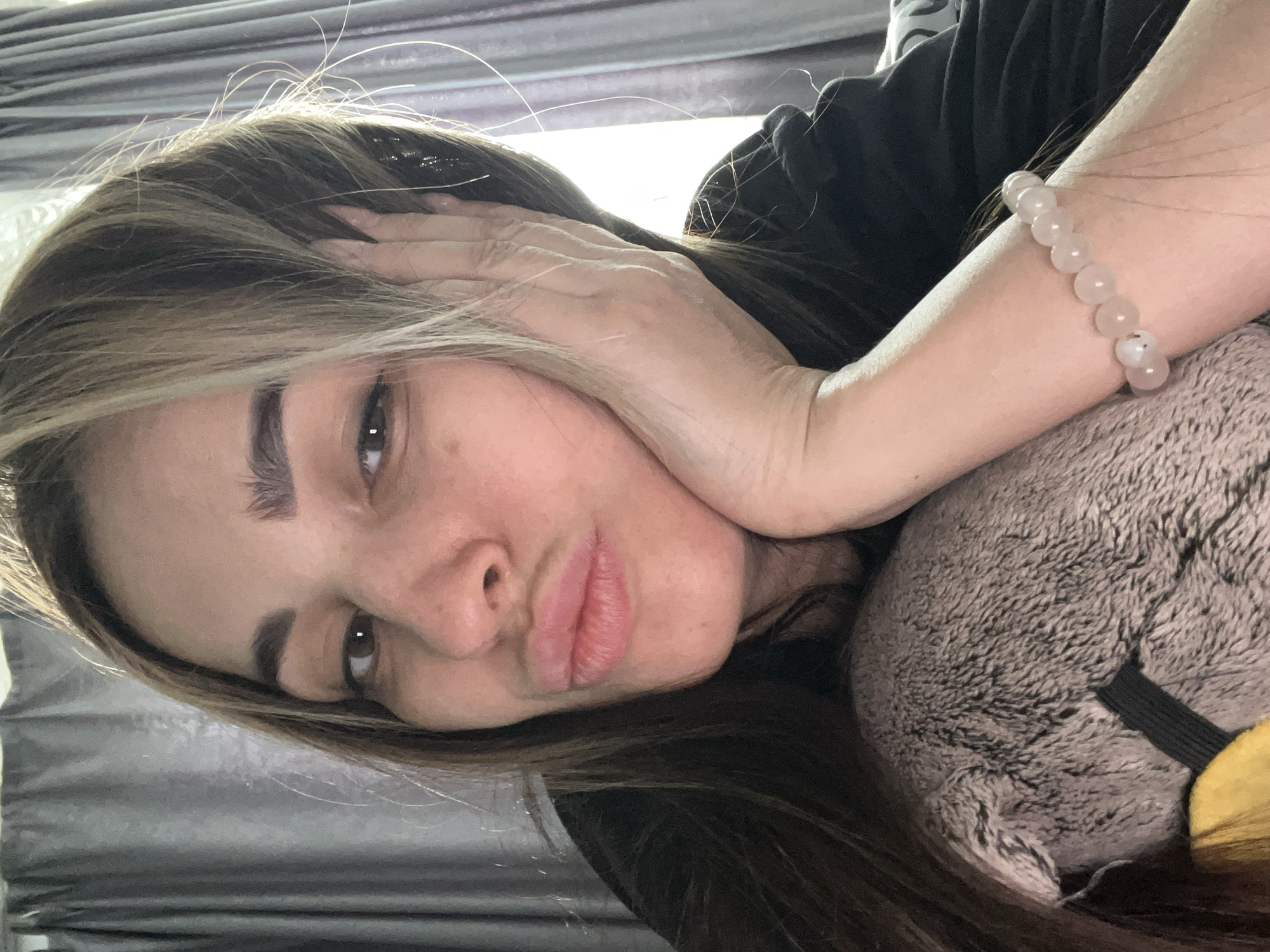 A woman is lying down, her head resting on a plush toy. She has light brown hair with blonde highlights, and is wearing a black top. Her left arm is bent with her hand near her face, and she is wearing a beaded bracelet on her left wrist. The background appears to be a gray curtain or wall. The location is unspecified.