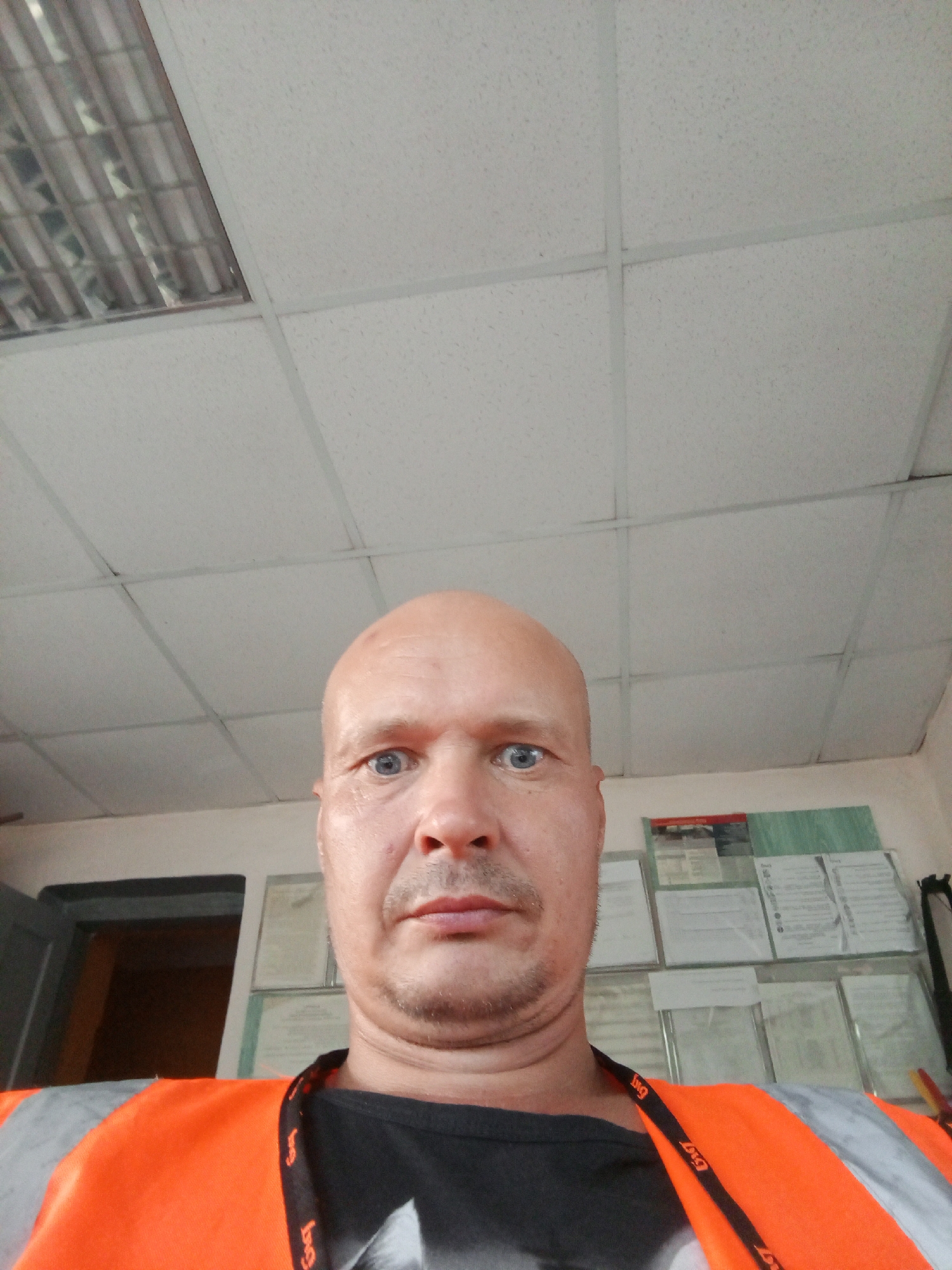 This is a selfie taken indoors. The subject is a bald, middle-aged man with blue eyes, wearing an orange safety vest over a black t-shirt and a black lanyard. The background shows a drop ceiling with fluorescent lighting, and a wall with several framed documents or notices. The overall impression is an office or work environment. The exact location in Kamensk-Uralsky, Russia cannot be determined from the image alone.