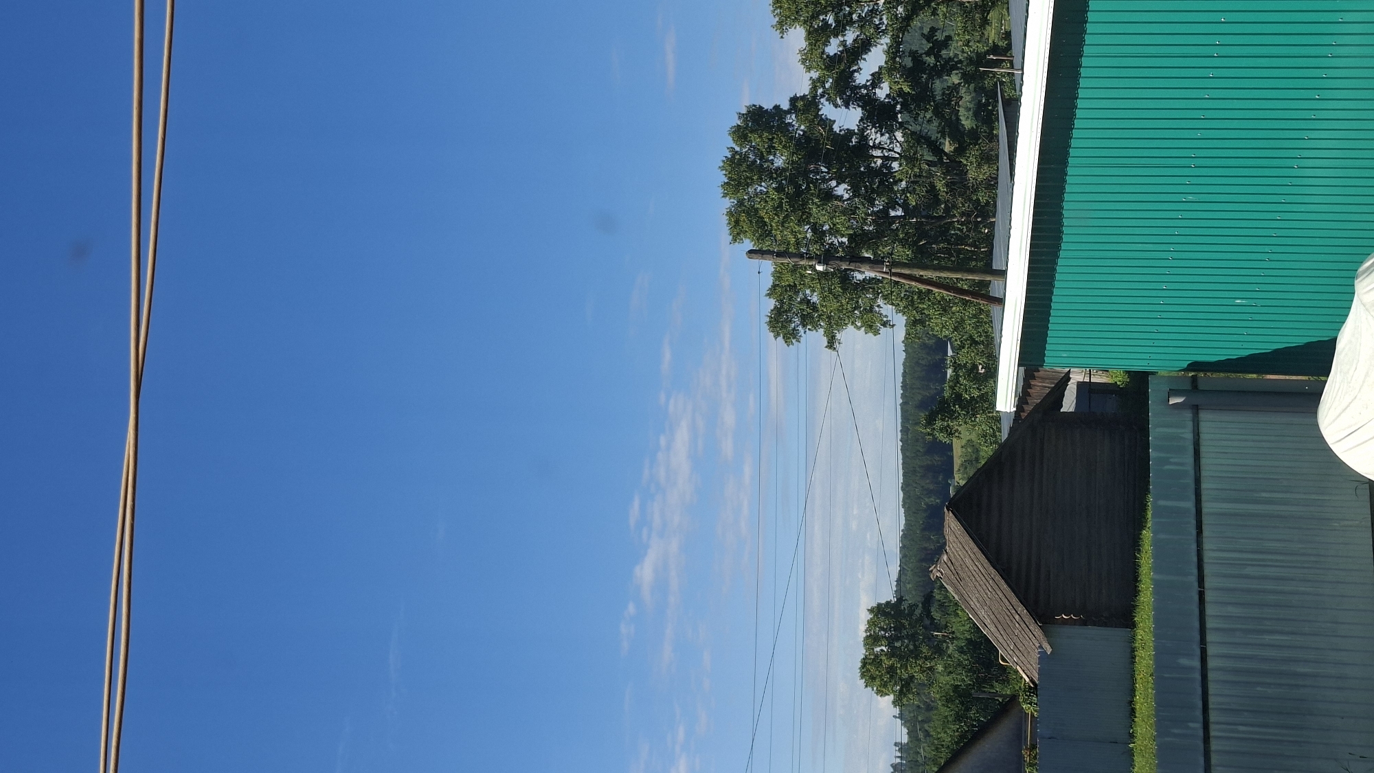 This image depicts a rural scene in Большой Букор, Russia, under a clear blue sky with scattered white clouds. The orientation of the photo is sideways. In the foreground on the left, two thin, brown wires are suspended against the sky.
Dominating the right side of the frame is a bright green corrugated metal roof, likely part of a building or shed. To its left, a smaller, darker corrugated roof is visible, and below that, a section of a fence or wall. Further to the left, a wooden utility pole stands with insulators, supporting electrical wires that stretch across the middle ground.
Behind the structures and utility pole, a lush green landscape unfolds, characterized by rolling hills covered in dense trees. The time of day appears to be daytime, judging by the bright sunlight and clear sky, suggesting fair weather. The overall impression is one of a tranquil, natural setting with elements of human habitation. No people are visible in the image, and there is no visible text.