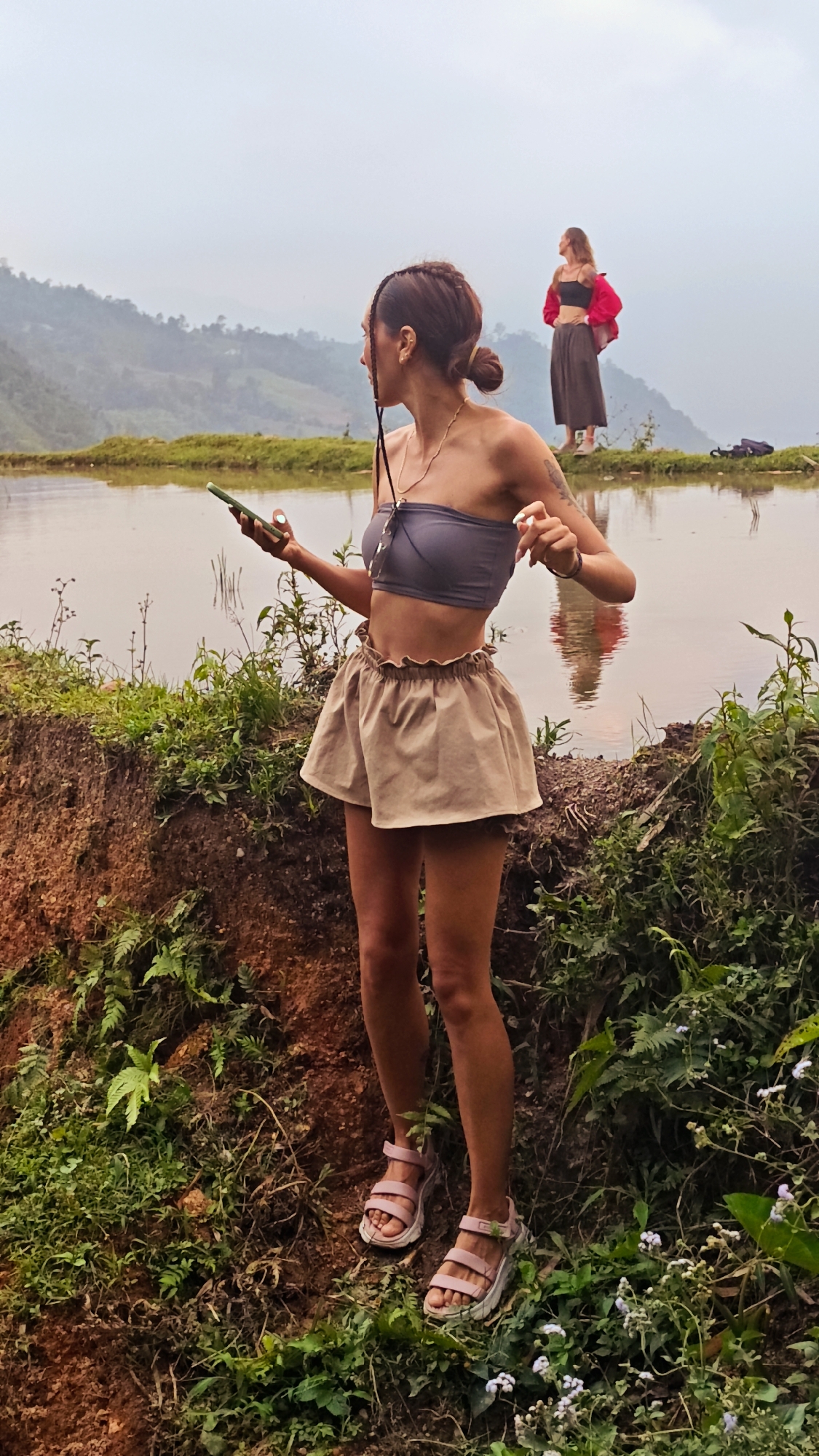 This outdoor, full-shot photograph depicts two women against a backdrop of a natural, mountainous landscape in Nậm Dịch Commune, Vietnam.
In the foreground, a woman with dark hair, pulled back into a bun with a side braid, stands on a muddy bank. She wears a grey strapless crop top, a light beige ruffled mini skirt, and light pink platform sandals. She holds a green smartphone in her left hand, and sunglasses hang from a necklace around her neck. A tattoo is visible on her right arm. Her gaze is directed towards the left, off-camera.
In the mid-ground, a body of calm water, likely a rice paddy or pond, stretches across the scene, reflecting the cloudy sky and a partial reflection of the background figure. Across this water, on a raised bank, a second woman with lighter hair stands. She is dressed in a black top, dark wide-leg pants, and a red jacket draped over her shoulders, and is looking out at the distant scenery. A dark bag or backpack lies on the ground beside her.
The immediate surroundings feature lush green vegetation and ferns growing on the muddy banks. The background is dominated by a series of rolling hills and mountains, partially obscured by an overcast and hazy sky, suggesting either early morning mist or a cloudy afternoon. The overall mood is serene, depicting people enjoying a scenic, rural environment. No text is visible in the image.