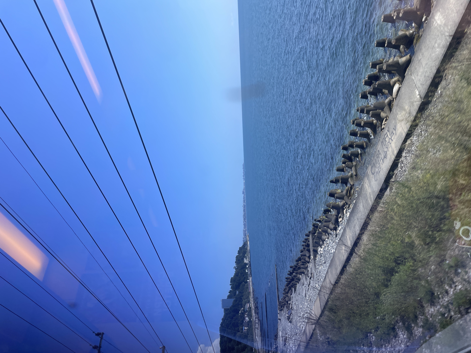 The image captures a view of the Black Sea coast in Sochi, Russia, as seen from a moving vehicle, likely a train or car, due to the streaks of light across the top of the frame and the slight motion blur. The scene is dominated by a clear, bright blue sky and the calm, deep blue waters of the sea. Along the shoreline, a concrete pathway runs parallel to the water, bordered by a rocky beach. A series of concrete tetrapod breakwaters are visible along the edge of the sea, designed to protect the coastline from erosion. In the distance, a green, tree-covered hillside rises, with buildings, including a tall, modern structure, nestled among the trees. Further along the coast, the urban landscape of Sochi stretches out, with more buildings visible on the horizon. The overall impression is one of a sunny day on the coast of a Russian resort city.