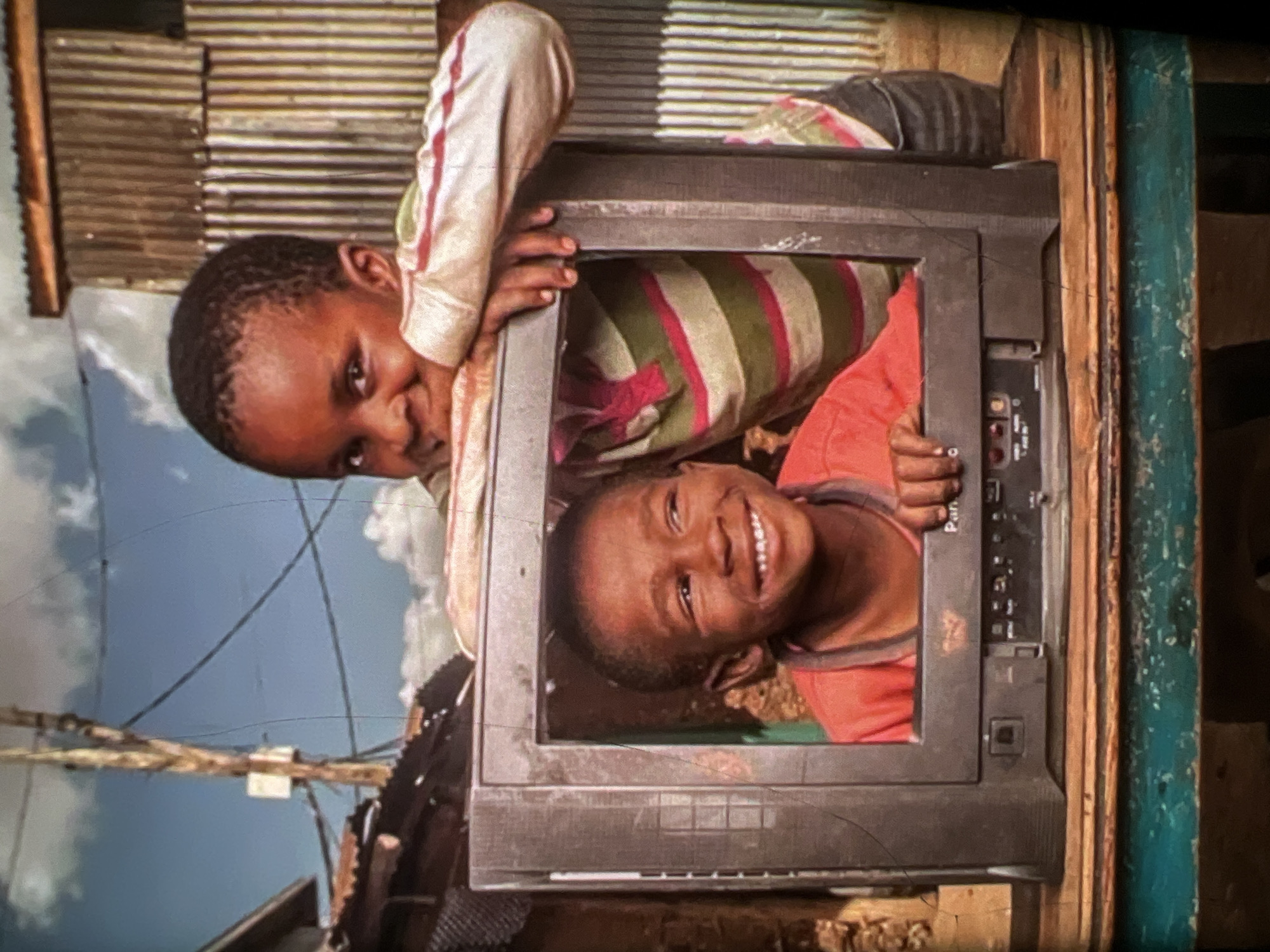 The image shows two young boys in what appears to be an outdoor setting in Lagos, Nigeria. In the foreground, a vintage television set is placed on a wooden surface, which is painted teal. A young boy, wearing an orange t-shirt, is positioned inside the television's frame, looking to his right with a wide smile. Behind him and slightly to his right, another boy, wearing a striped long-sleeved shirt with green, pink, and white stripes, leans over the television, resting his arms on its frame and looking towards the camera with a happy expression. The background features a corrugated metal structure, possibly a dwelling, and hints of other makeshift structures, suggesting an urban, possibly informal settlement. Overhead, a utility pole with wires and a cloudy blue sky are visible, further establishing an outdoor environment. The overall scene evokes a sense of joy and simple pleasure derived from shared moments and perhaps the novelty of the television.