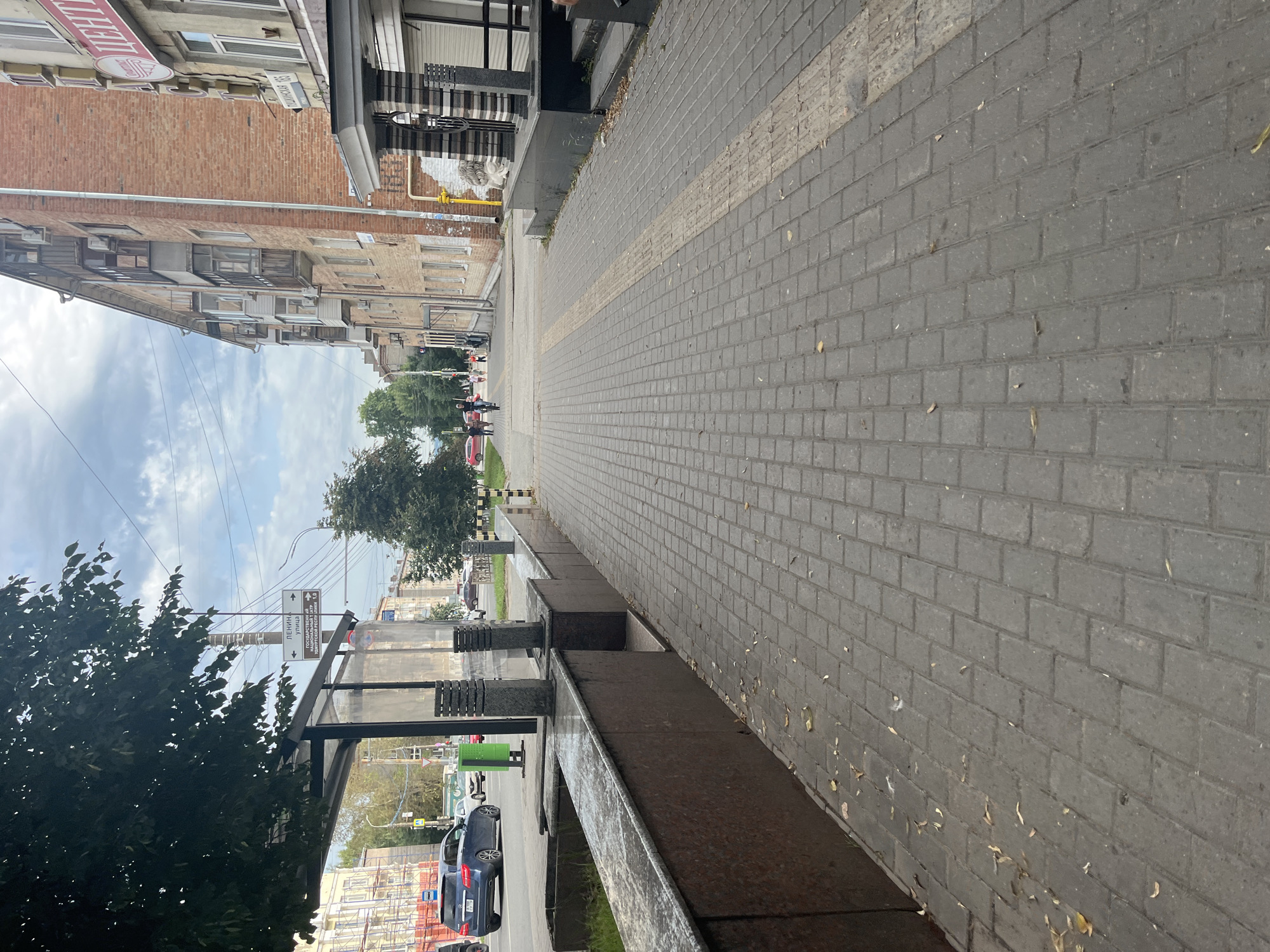 The image depicts a sidewalk on a cloudy day in Izhevsk, Russia. The sidewalk is paved with grey rectangular bricks and has a tactile paving strip running down its center. To the left, a blue car is parked near a bus stop with a green bin. Beyond the bus stop, there is a brick building with balconies and windows. To the right, another brick building features a store with an awning and the Russian word "ЦЕНТР" (Center) visible on its facade. Several pedestrians are walking on the sidewalk in the distance. The street is visible on the left with more cars. Trees and overhead wires are also present in the scene.