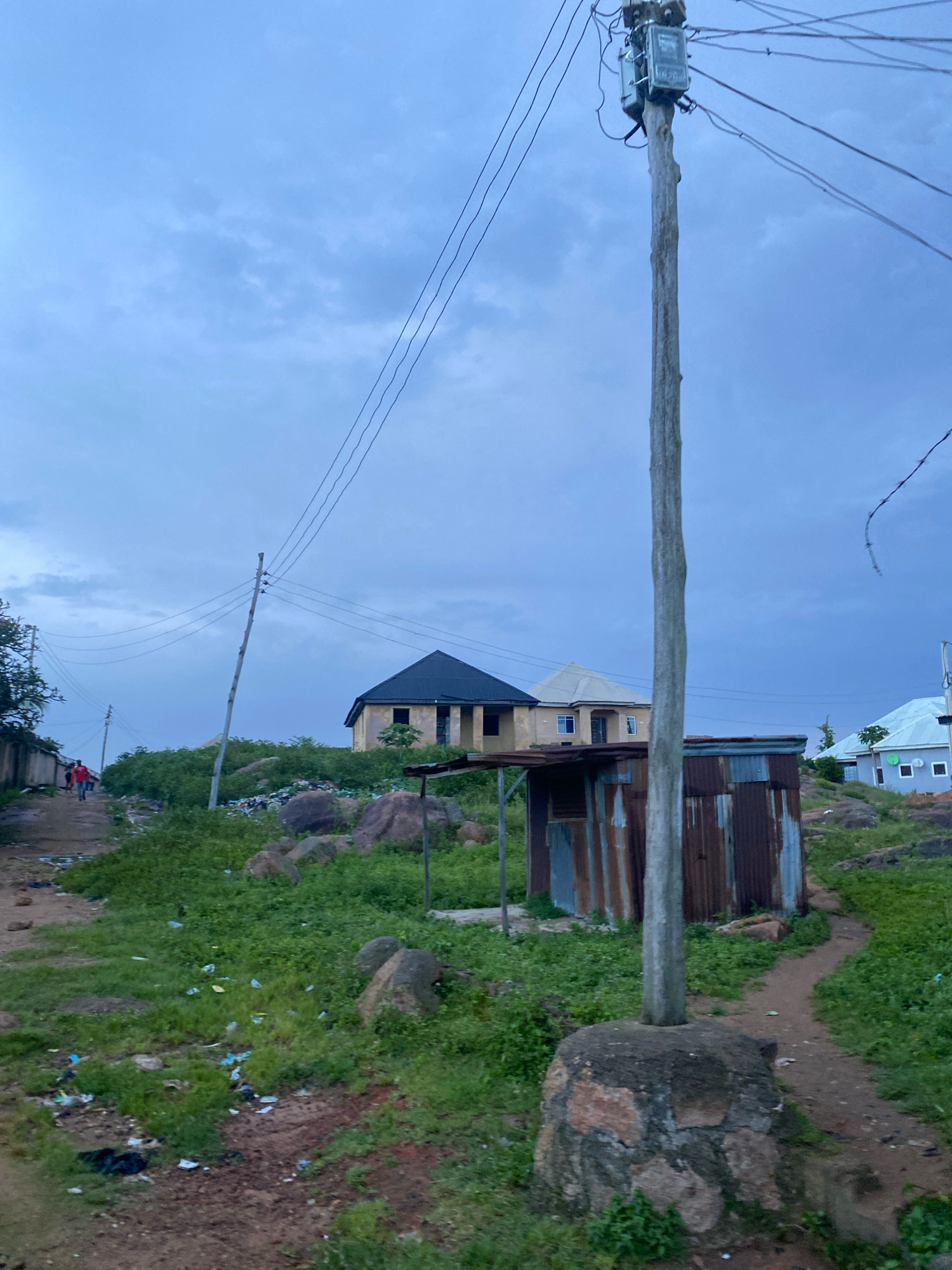 The image depicts a street scene in Jos, Nigeria, under a cloudy, overcast sky. A wooden utility pole with electrical meters and wires dominates the foreground, planted in a large, rough stone base. To the left of the pole, a path leads up a grassy incline littered with trash, including plastic bags and wrappers. Further up the hill, two unfinished houses are visible; one with a dark pitched roof and pale walls, and another with a lighter, similarly shaped roof. A corrugated metal structure, possibly a shelter or small shop, stands near the base of the utility pole. In the background, on the left side of the image, a dirt road curves away, with two figures walking away from the camera. Beyond the dirt road, more houses are visible, some with light-colored walls and pitched roofs. The landscape is rocky and covered in lush green grass and bushes, with scattered boulders throughout.