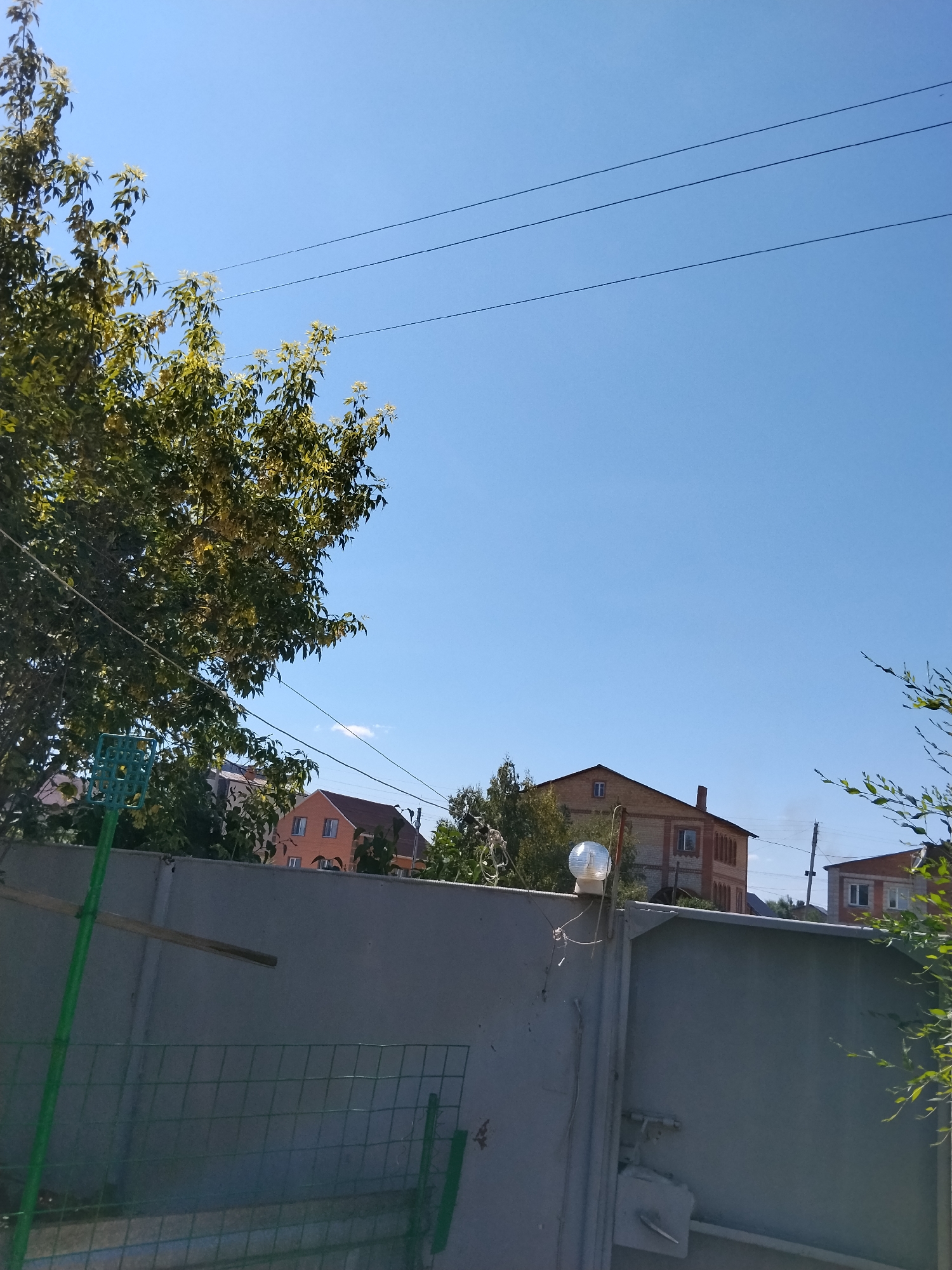 The image depicts an outdoor scene under a clear blue sky, likely in Kokshetau, Kazakhstan, given the provided context. The foreground is dominated by a tall, grey, corrugated metal gate, which appears to be part of a fence. Attached to the top of the gate, on the left side, is a small, round, white light fixture. To the left of the gate, a green metal pole extends upwards, supporting a green, square-shaped object with a grid pattern, possibly a fly swatter or a similar tool. Further left, a portion of a lush green tree with broad leaves is visible.
Behind the gate, several houses are partially visible. The closest house on the left is light orange with white window frames. To its right, a larger, brick house with red brick accents and white window frames is prominent, featuring a distinctive gable roof. In the distance, more structures and trees can be seen. Three dark power lines are stretched across the upper portion of the frame against the sky. The overall impression is of a residential area on a bright, sunny day. There are no people or animals visible in the image.