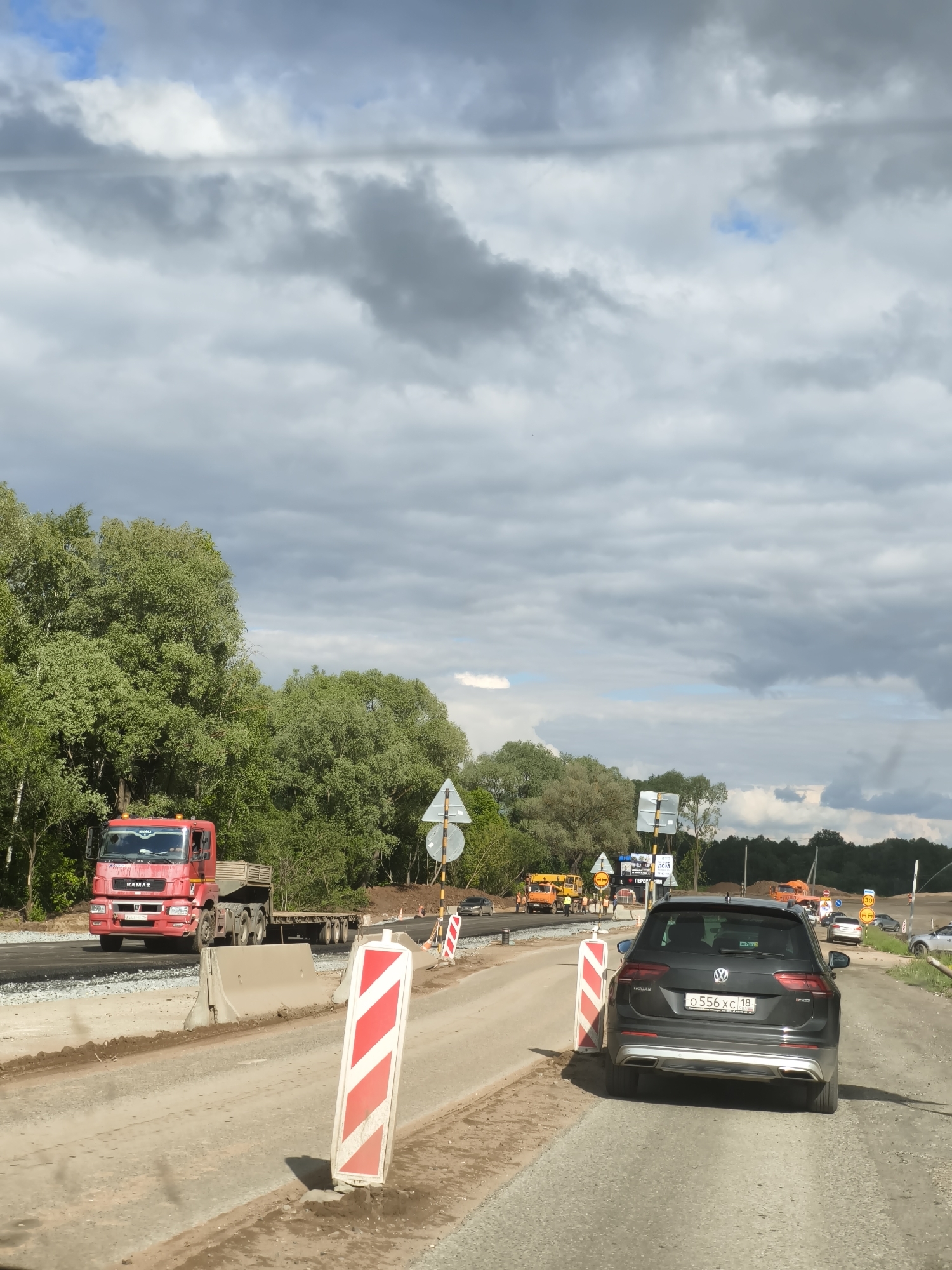 The image depicts a road construction site in Kabanikha, Russia, under a cloudy sky with glimpses of blue. The foreground features a temporary dirt road section flanked by red and white striped warning markers and concrete barriers.
On the left, a large red KAMAZ truck with a flatbed trailer is driving on a newly laid asphalt section of the road. The truck has "KAMAZ" branding visible on its cab and grille, and a Russian license plate. A person is visible in the driver's seat. Adjacent to the road, dense green trees line the landscape.
In the mid-ground, beyond the barriers, construction activity is evident with yellow heavy machinery and several workers in orange safety vests. Various road signs are scattered, including one indicating a "30" speed limit and others with Russian text, such as "ПЕРЕЕЗД" (crossing) and "ЛЮМ".
In the right foreground, a dark grey Volkswagen Tiguan SUV is parked or moving slowly, facing away from the viewer. The vehicle displays "Volkswagen Tiguan" badging and a clear Russian license plate "o 556 xc 18". The sky above is largely covered by grey clouds, though some patches of clear blue sky are visible. The overall scene indicates daytime with ongoing road work.
