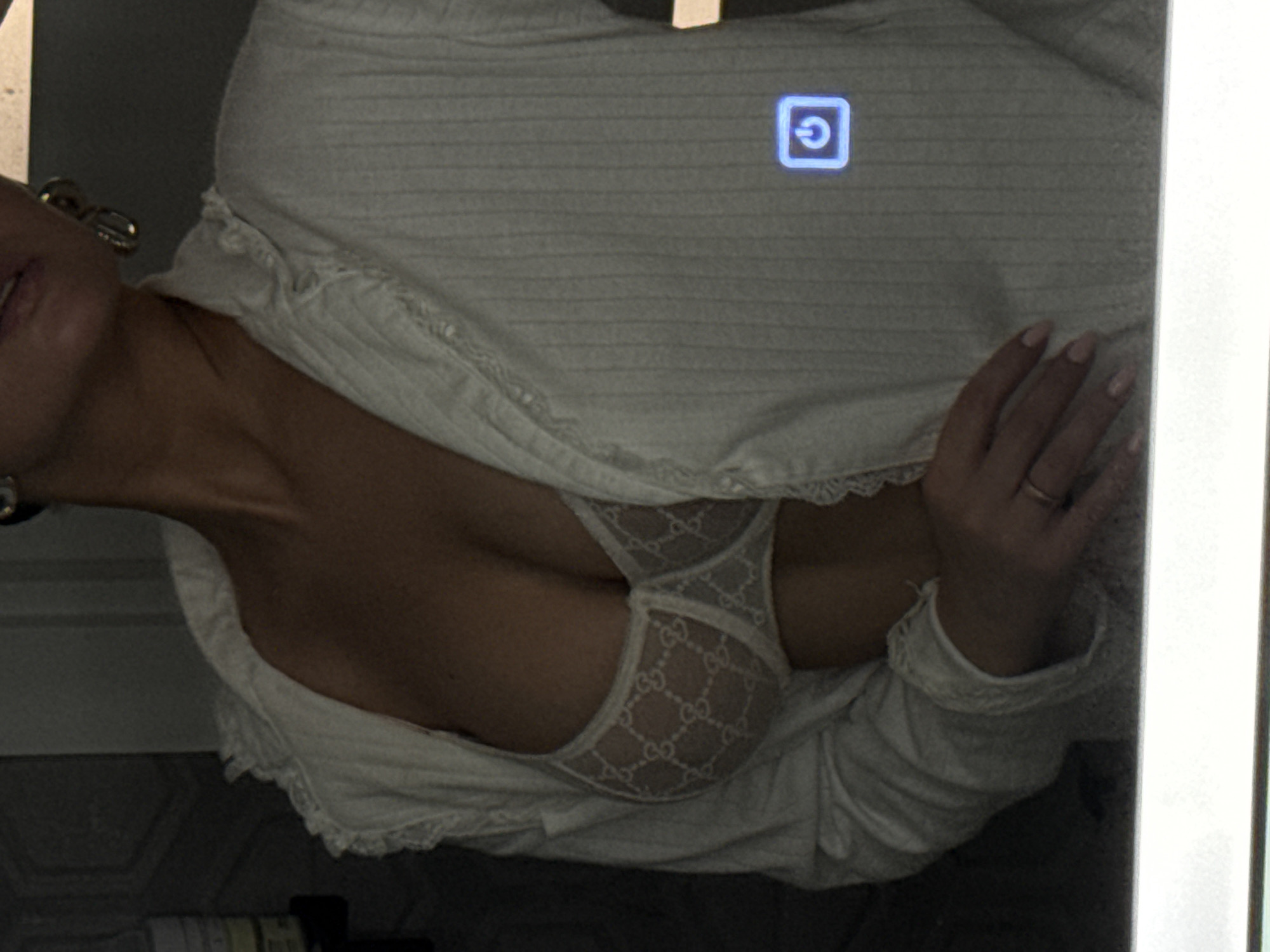 The image shows a woman partially visible, lying down. She is wearing a white, lace-trimmed top or robe over a white bra with a "Gucci" pattern. Her right hand is raised near a reflective surface with a power button icon. The background is dark and indistinct. There is no information available to determine the location as Chaikovsky, Russia, or any other specific geographic location.