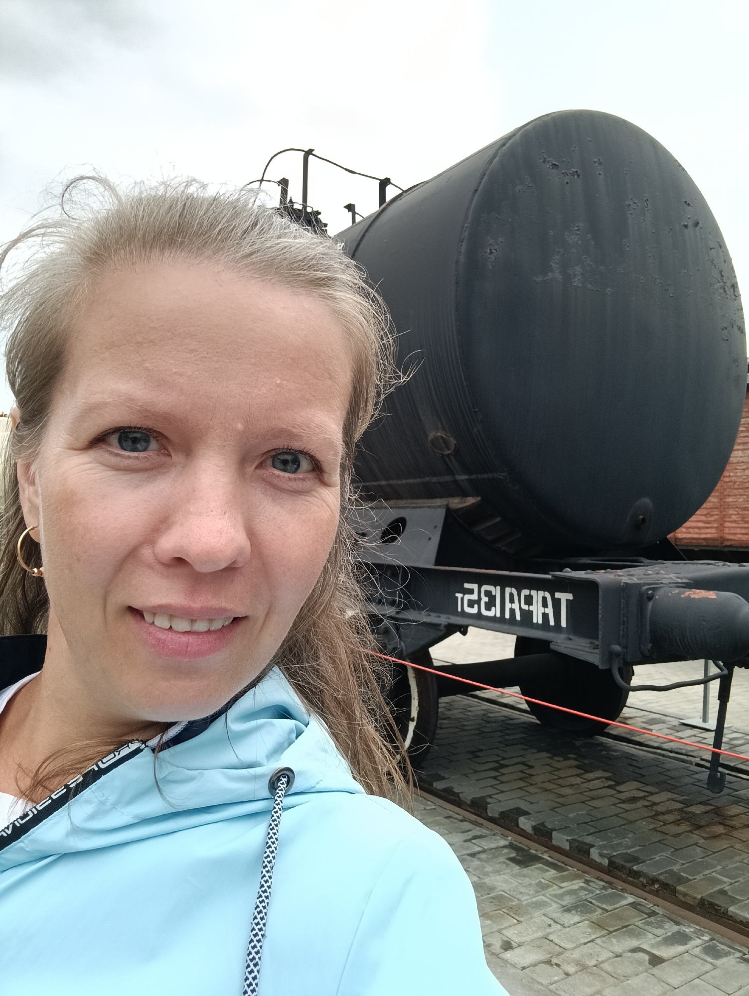 A woman with blonde hair and blue eyes is taking a selfie outdoors. She is smiling and wearing a light blue jacket with a black and white patterned trim around the hood. Behind her, a large black cylindrical tank car is visible, resting on railway tracks. The tank car has some rust spots and a white inscription that appears to be reversed, reading "T2E1A9AT". The ground is paved with cobblestones, and the sky is overcast, suggesting it is a cloudy day. The woman is positioned in the foreground, and the tank car dominates the background, indicating an industrial or historical site, possibly a museum given the context of Verkhnyaya Pyshma, Russia, known for its military equipment museum. The woman's expression is cheerful.