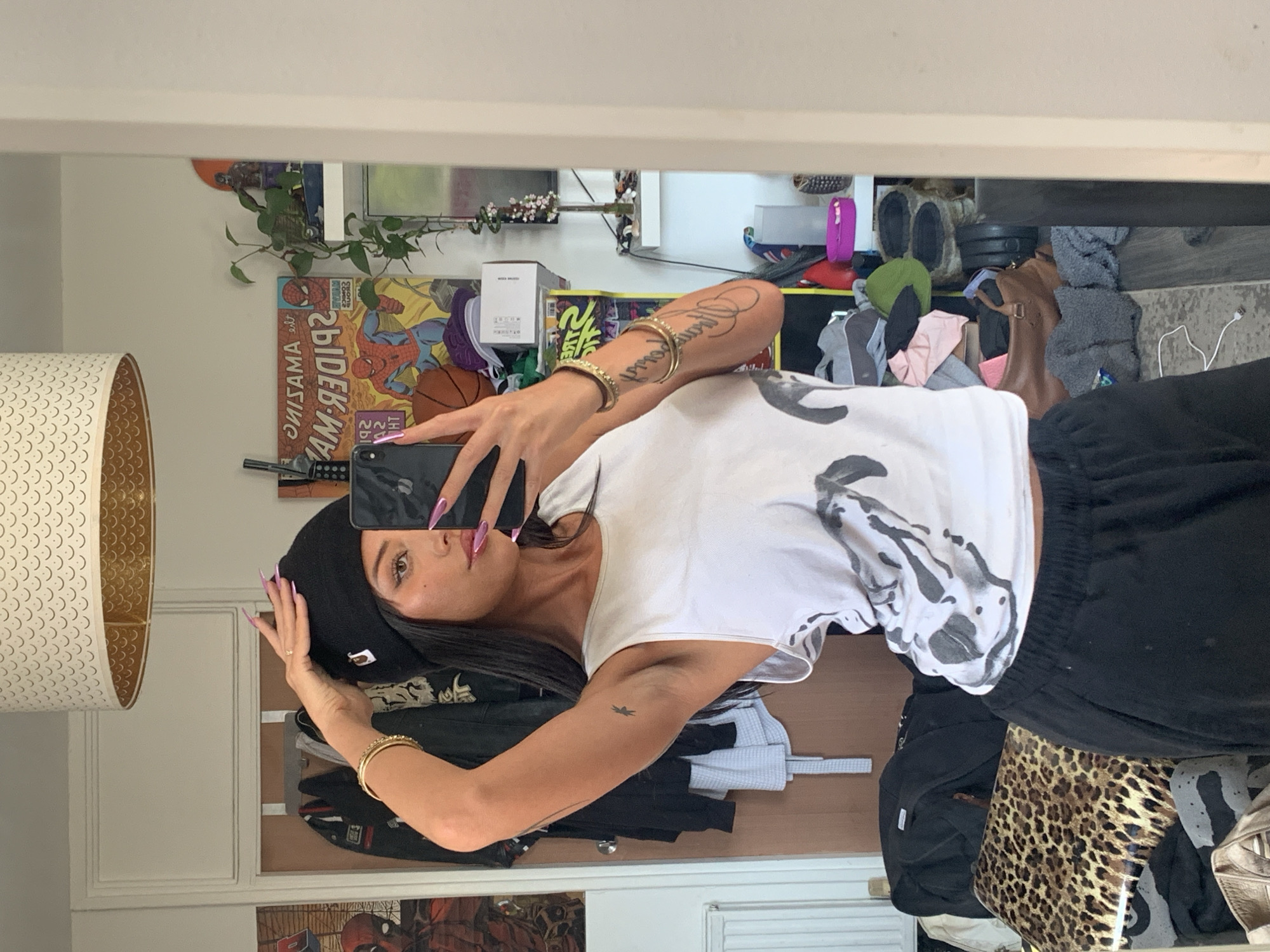 This is a mirror selfie of a young woman taken indoors. She is positioned centrally, holding a dark-colored smartphone with her right hand to capture her reflection. Her long, dark hair frames her face, and she has brown eyes, styled eyebrows, and wears makeup. She is wearing a black beanie with a small white logo, a white sleeveless top with an abstract dark design, and black sweatpants. Several gold-colored bracelets are on her right wrist, and she has visible tattoos, including script on her right forearm and a small bird-like symbol on her left bicep. Her nails are long and painted a bright pink/purple hue.
The background, seen in the mirror's reflection, reveals a personal indoor setting, likely a room or bedroom. To the left (viewer's right), a large lamp with a patterned, crescent-shaped shade is visible. Above the woman's head, a "The Amazing Spider-Man" comic book cover is prominently displayed, showing Spider-Man and a basketball, with the reversed text "AMAZING SPIDER-MAN" and numbers "192" visible. A basketball and several plants, including a trailing vine, are also in this area.
Further back, the room appears to have storage units or shelves filled with various items. These include piles of clothes in different colors (pink, grey, black), a brown bag, a green beanie, and some footwear. A distinctive leopard print item is visible near the bottom right of the reflection. White boxes and a white power cord are also present, indicating a lived-in and somewhat cluttered space. The overall ambiance is casual and personal.