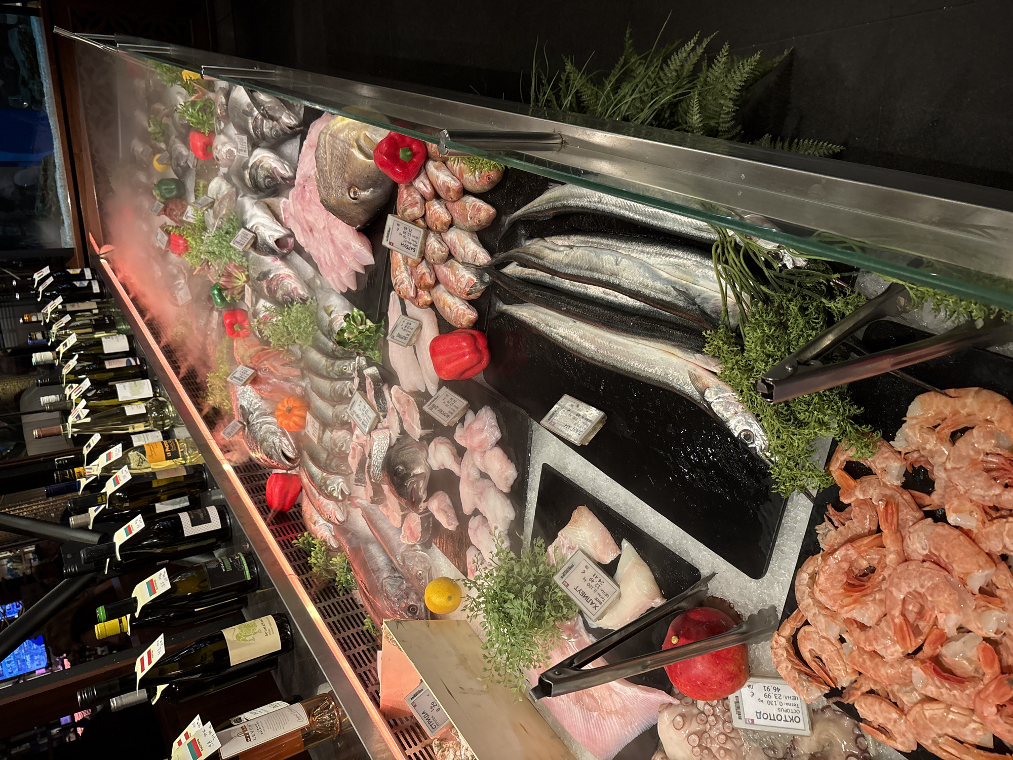 This image shows a display case of seafood and wine in a store, likely in Sofia, Bulgaria, based on the Cyrillic text on price tags. The display includes various types of fish, such as sea bass, red mullet, and possibly needlefish, alongside shrimp, octopus, and halibut fillet. Some red peppers and sprigs of greenery are used as decoration. Bottles of wine are visible on the left side of the image. The seafood appears to be on ice within the refrigerated display case. Tongs are available for customers to select the desired seafood. Price tags with Cyrillic text indicating weight and price are positioned beside the seafood items.