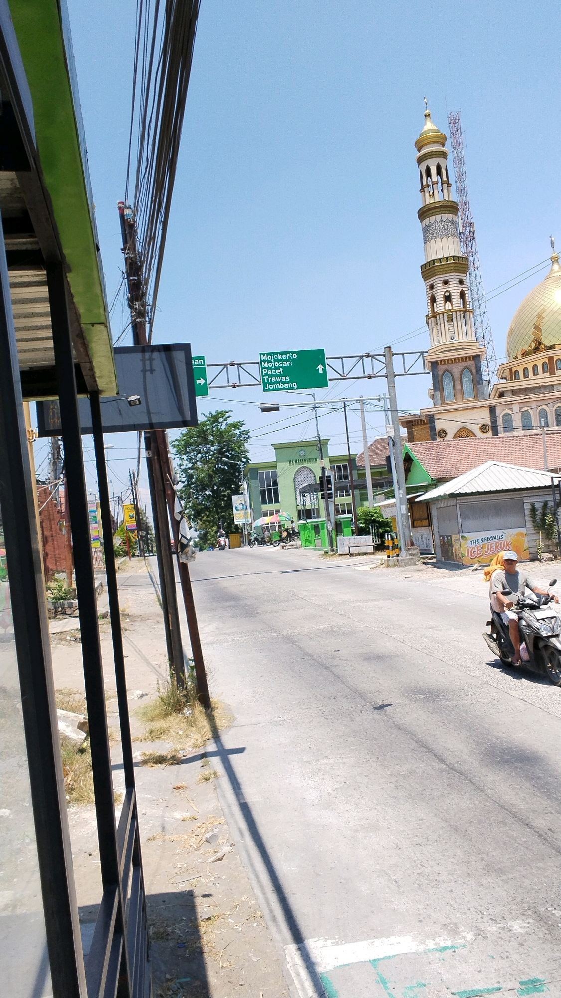 The image depicts a street scene in Pulorejo, Indonesia, on a bright, sunny day with a clear blue sky. In the foreground, a portion of a black metal structure with green trim is visible on the left, casting a shadow on the ground. The street itself is paved and appears to be a two-lane road.
Dominating the mid-ground and background is a large, ornate mosque with a golden dome and a tall, slender minaret topped with gold. The mosque is colored in light brown and cream with intricate architectural details. Adjacent to the mosque, on the right side of the street, is a small building with a white roof, advertising "THE SPECIALIS ICE JERUK" (Orange Ice).
A green directional signpost is suspended over the road, indicating "Mojokerto," "Milosari," "Pacet," "Trawas," and "Jombang" with arrows pointing left and straight ahead. Several green buildings with modern architecture are visible on the left side of the street, one of which has "MA NAFI'UDDIN" written on it. Various wires and utility poles are scattered throughout the scene.
In the right lane of the road, a person wearing a yellow headscarf and a grey shirt is riding a black motorcycle, heading away from the camera. Other people and a few motorcycles are seen further down the street, near the green buildings, suggesting a moderately busy area. The environment appears to be a mix of urban infrastructure and some dry vegetation along the roadside.