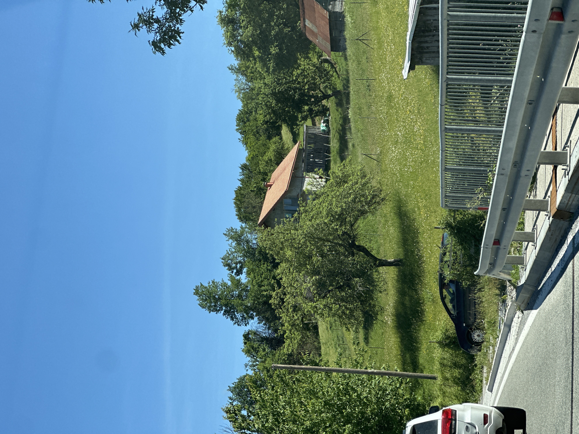This is a photograph taken in the city of Jabuka, Montenegro, on a clear, sunny day. The image captures a rural landscape with lush green hills and trees under a bright blue sky. In the foreground, a modern white SUV is partially visible on the left side of a paved road, suggesting the photograph was taken from inside another vehicle. To the right, a metallic guardrail lines the edge of what appears to be a bridge or elevated walkway, offering a view of the scenery beyond.
Further into the landscape, a dark-colored car is seen parked precariously on the grassy verge, partially obscured by overgrown vegetation. Behind the car, a gentle slope leads up to a modest house with a red-tiled roof. The house has a simple wooden porch or extension. Further to the right, a smaller, rustic wooden structure with a rusty metal roof is also visible. The hills are covered in vibrant green grass dotted with small white wildflowers, indicating a flourishing natural environment. A wooden utility pole stands near the left edge of the frame, and faint fencing can be observed in the midground, delineating the fields. The overall impression is one of a serene, possibly slightly neglected, rural setting on a warm day.
