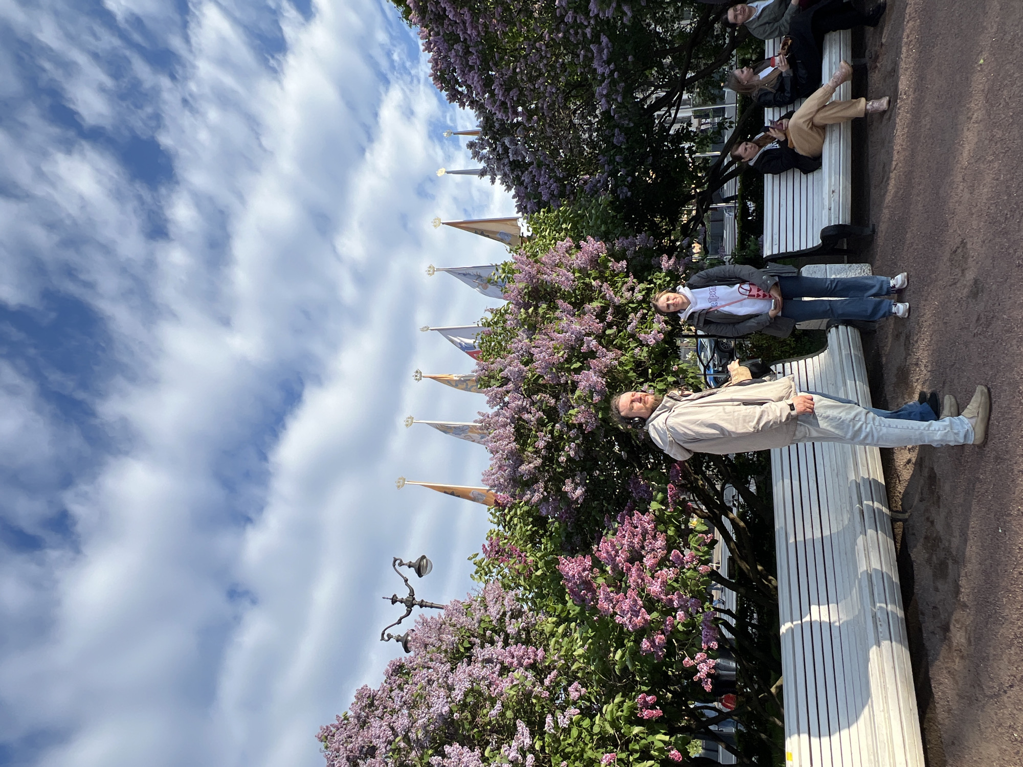 The image shows a partially sunny day in Saint Petersburg, Russia. A group of people are relaxing on a white bench near a park or garden filled with blooming lilac bushes. Flags adorned with royal emblems and spires can be seen in the background, which is possibly indicative of Peterhof or other royal residences. The people are interacting with each other, relaxing, and enjoying the weather. A street light is located adjacent to the bench. The sky is blue with scattered clouds.