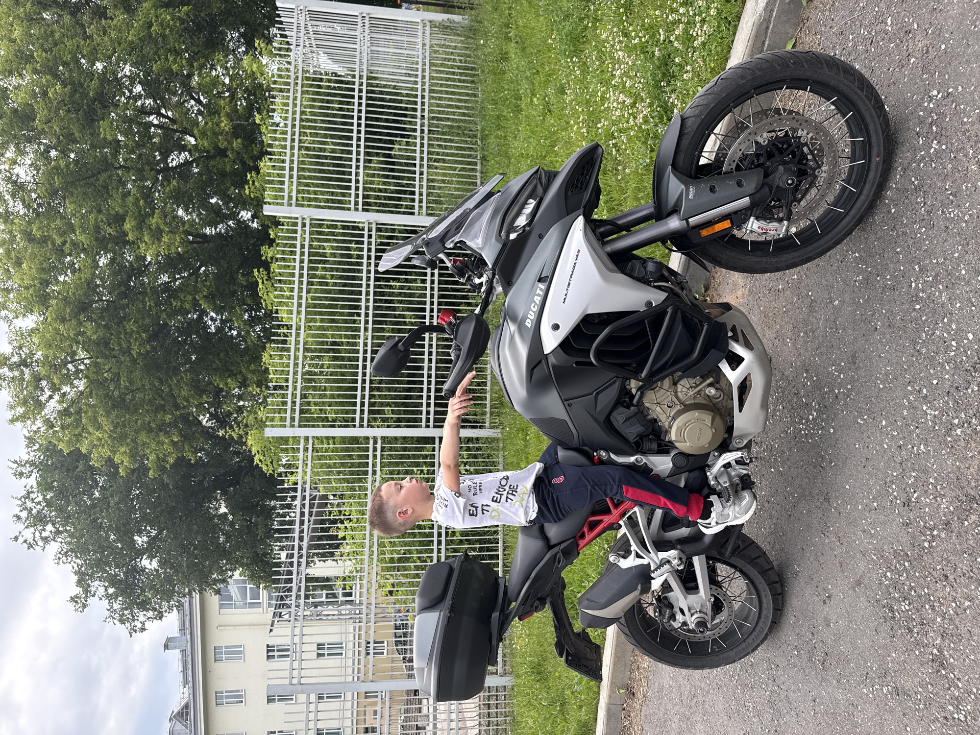 A young boy is sitting on a Ducati Multistrada V4S motorcycle. He is wearing a white t-shirt with text and dark blue pants with red stripes. The motorcycle is grey and silver. The boy is pointing his left hand upwards. In the background, there is a white fence, green grass with small white flowers, and a building. The sky is overcast. The scene appears to be outdoors, possibly a street or parking area, given the asphalt and curb.
The text on the boy's t-shirt is partially visible and includes "ENJOY THE". The motorcycle has "DUCATI" and "MULTISTRADA V4S" written on it. The overall atmosphere seems casual and possibly a moment of play or imagination.
The image is oriented vertically, and the scene is captured in daylight under cloudy conditions. The location context is Votkinsk, Russia.