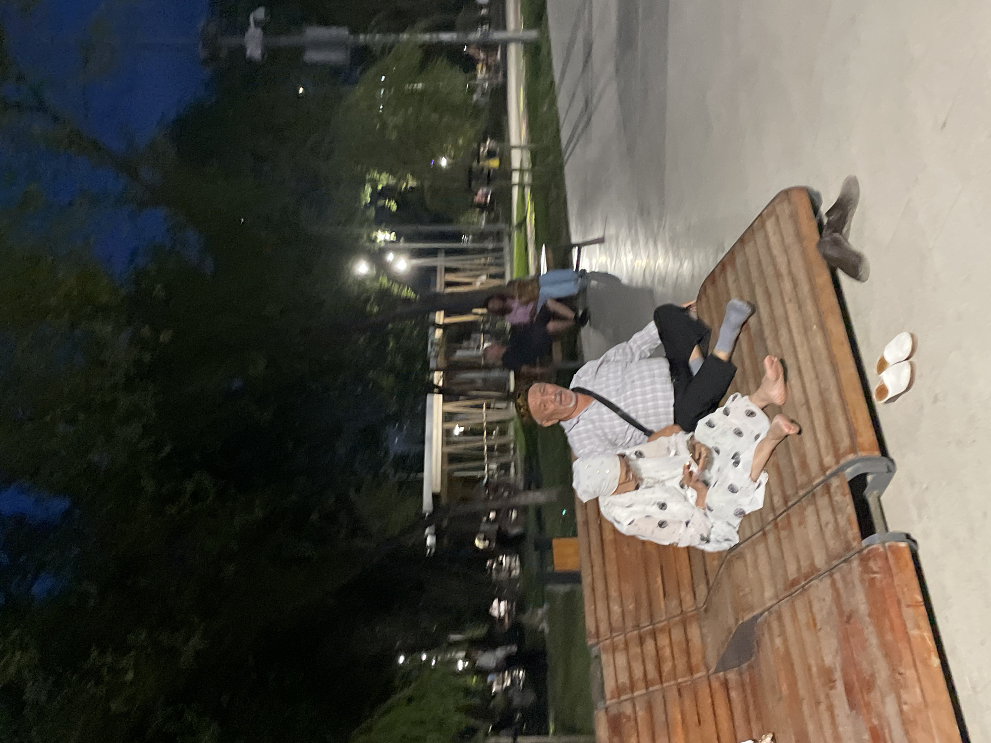 A man and a woman are sitting on a wooden bench in a park at dusk. The man, wearing a checkered shirt and a cap, is looking to the right. The woman, in a white patterned dress and headscarf, is looking down at her phone. Their shoes are placed on the ground near the bench. In the background, trees, lampposts, and other park-goers are visible. The scene appears to be in Almaty, Kazakhstan, likely in a public park such as Panfilov Park or the Botanical Garden, given the ambiance and typical park structures.
