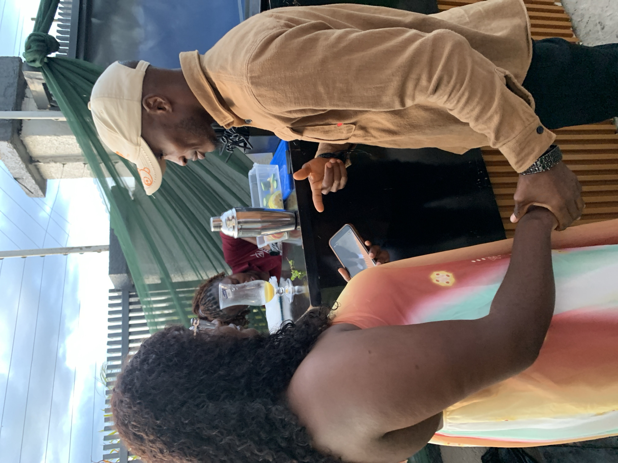 The image shows a man and a woman standing at a bar with a green tarp in the background. The man is wearing a tan shirt and a tan baseball cap. He is looking at the woman and gesturing with his right hand. The woman is wearing a colorful dress and holding a phone in her left hand. There is a cocktail shaker and a glass of orange liquid on the bar. The scene appears to be outdoors, possibly in Gura Top, Nigeria, given the context. The sky is cloudy.