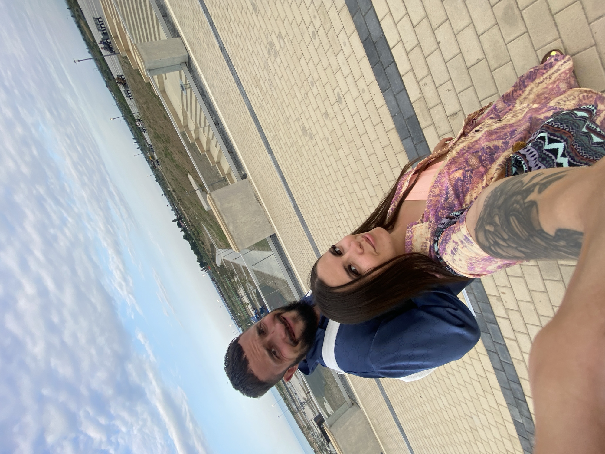 A man and a woman are taking a selfie outdoors, with the city of Arablyar, Russia visible in the background. The man, with a beard and a short haircut, is wearing a blue hoodie. The woman, with long dark hair, is wearing a pink top and a colorful patterned dress. They are standing on a paved area with a tiled walkway and stairs. The sky is partly cloudy. The location appears to be a public space or park, possibly with a view of the sea, given the distant coastline and the open expanse.