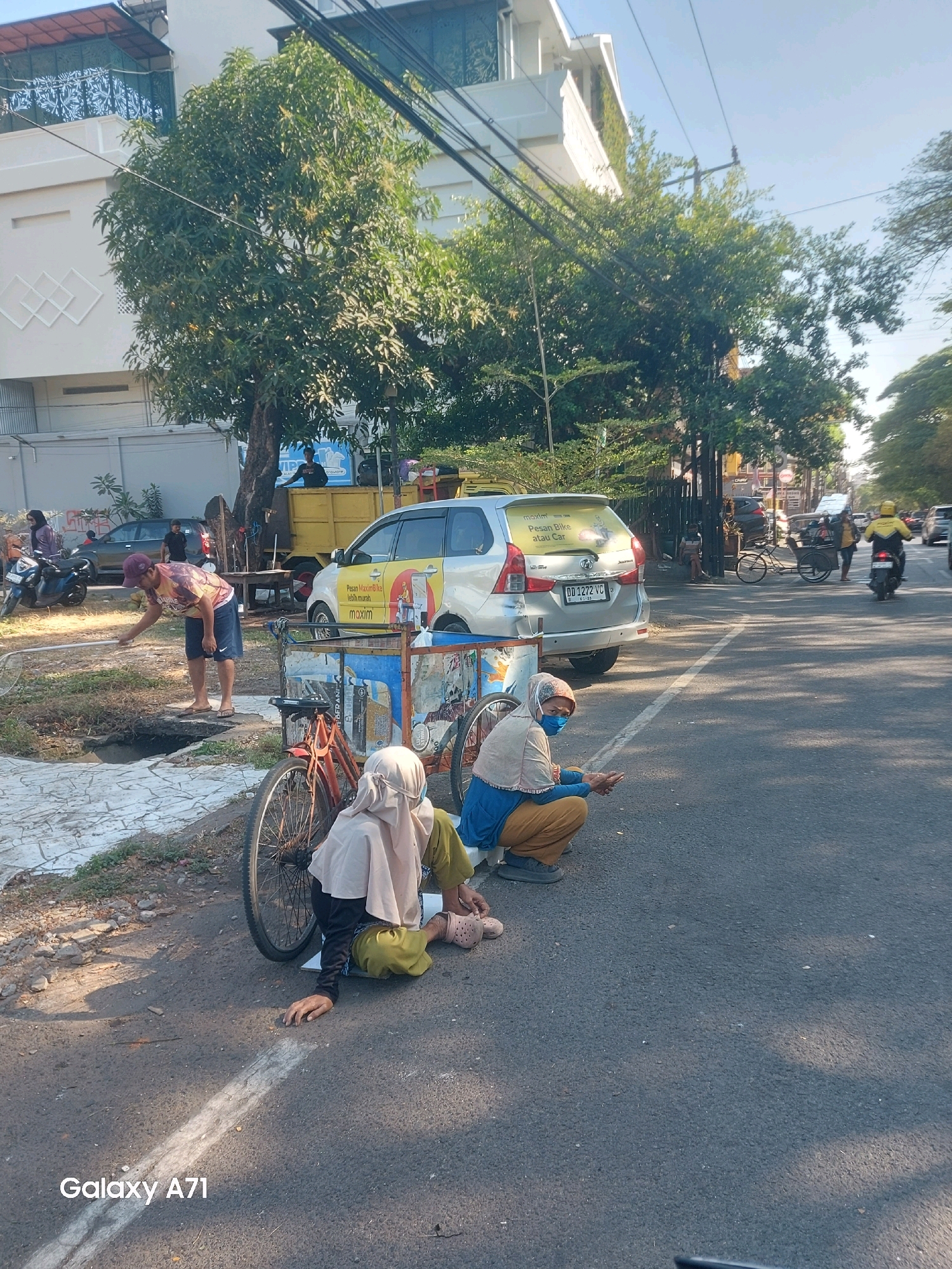 This image captures a street scene in Makassar, Indonesia, under a bright, clear sky. In the foreground, two women are seated on the asphalt by the side of the road, near a parked bicycle. Both are wearing hijabs; one in a light tan and the other in a darker tan. The woman on the left wears a black top and a green outer garment, while the woman on the right is dressed in a blue top and brown pants. A red bicycle is partially visible, its front wheel obscuring the woman on the left.
Behind them, a silver car is parked, bearing a sticker with Indonesian text. A yellow truck is also present, with a person standing in its bed. Further back, trees line the street, casting dappled shadows. A man in a maroon cap and blue shorts is seen in the middle ground, appearing to be cleaning or tending to something near a drain. In the distance, traffic, including motorcycles and a bicycle rickshaw, moves along the street. The overall impression is one of a typical urban environment with everyday activities unfolding.
