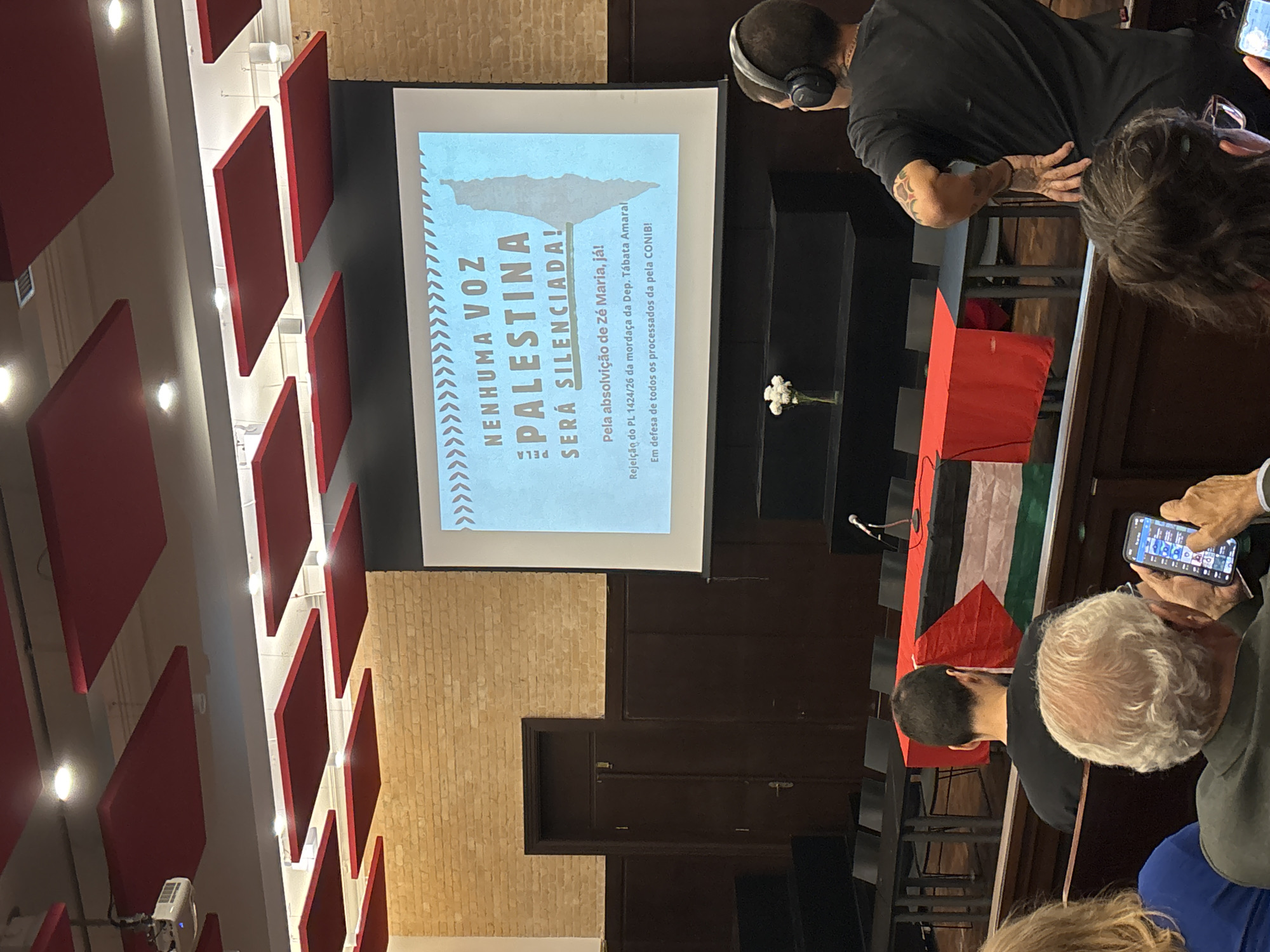 Here is a detailed description of the image:
**Scene:** The image captures a scene inside a meeting room or auditorium in what appears to be São Paulo, Brazil, given the Portuguese text on the screen. The room is set up for a presentation or discussion.
**People:** Several people are present, although the image only shows them from the shoulders up or partially. There is a man wearing headphones, another man with visible tattoos on his arm, and several others in the audience. One person is holding and looking at a smartphone.
**Subjects:** The main subject is a projected screen displaying a message in Portuguese about Palestine. The text translates to "No voice of Palestine will be silenced!" It also mentions something about the absolution of "Zé Maria" and the rejection of a PL (likely a legislative project) concerning freedom of speech. The text also makes reference to the CONIB, the Confederacao Israelita do Brasil, which translates to the Israelite Confederation of Brazil.
**Objects:** In addition to the screen, the room contains:
- A stage with a podium or table draped with a Palestinian flag.
- A microphone on the podium.
- A small bouquet of white flowers.
- Chairs arranged for the audience.
- Ceiling panels, some of which are red.
**Actions/Interactions:** The people are generally in listening mode, with one person actively using their phone. The event seems to be centered around advocating for Palestine and possibly protesting restrictions on free speech.
**Environment:** The room has a modern feel with architectural details such as exposed brick and geometric paneling on the ceiling. The lighting is moderate.
**Overall impression:** It's a snapshot of an event that combines political activism and advocacy for Palestinian rights, possibly reacting to local political dynamics and related to freedom of speech issues in Brazil.