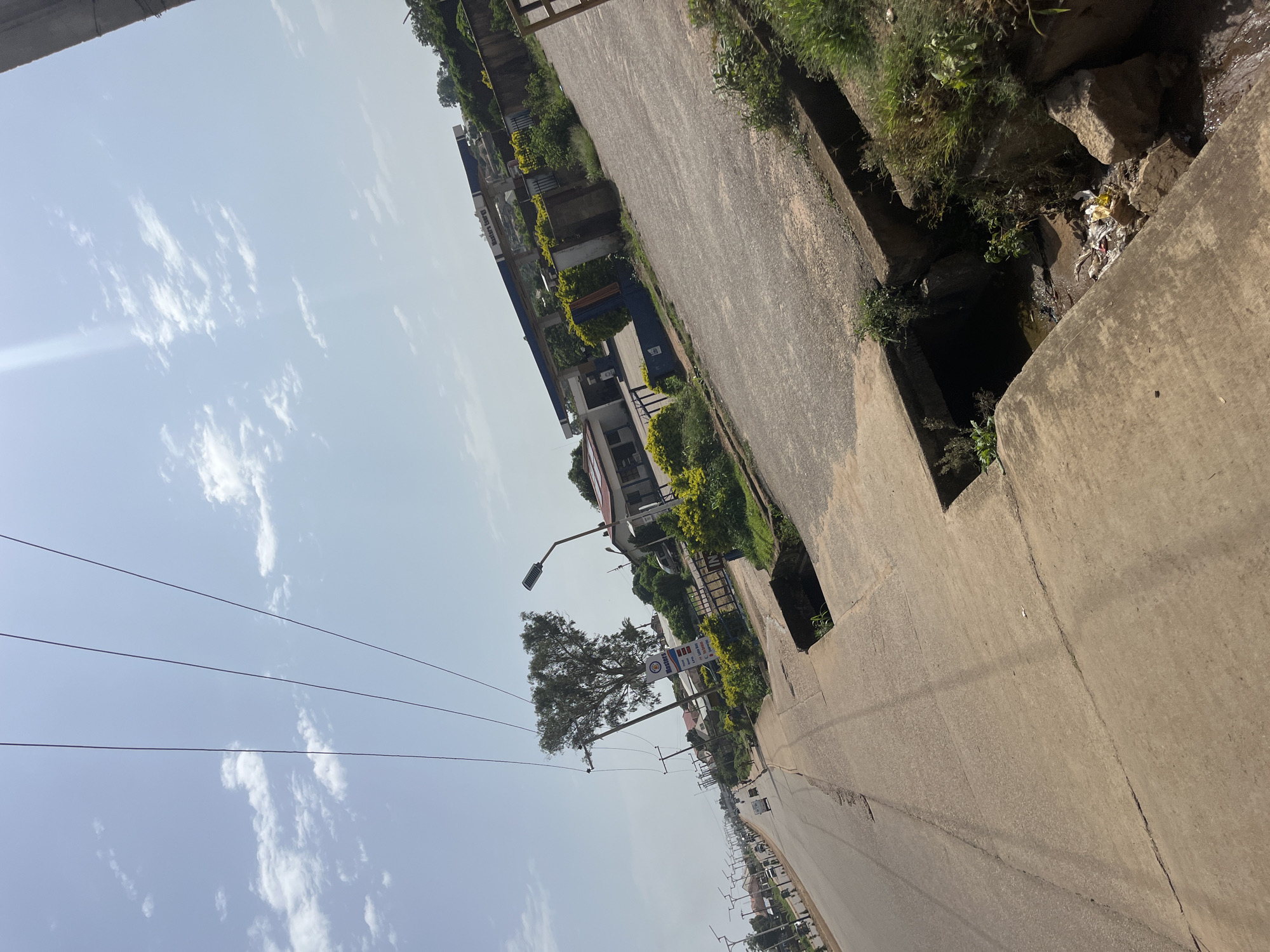 The image shows a view of a road in Bukuru, Nigeria, on a bright, cloudy day. The road runs from the foreground into the distance, with a concrete sidewalk on the right. Along the sidewalk, there is a drainage ditch filled with some debris and overgrown vegetation. A large tree with sparse foliage stands near the road, accompanied by a utility pole with multiple wires.
Across the road, a gas station is visible, identified by its blue and white canopy structure with the words "Masters Energy" printed on it. Adjacent to the gas station are low-lying buildings and lush green bushes. Several streetlights are positioned along the side of the road. In the distance, more buildings and trees can be seen, suggesting a developed urban or semi-urban area. A few vehicles are present on the road, and a person can be seen walking on the sidewalk in the mid-ground. The sky is a light blue with scattered white clouds. The overall impression is of a typical street scene in a Nigerian city.