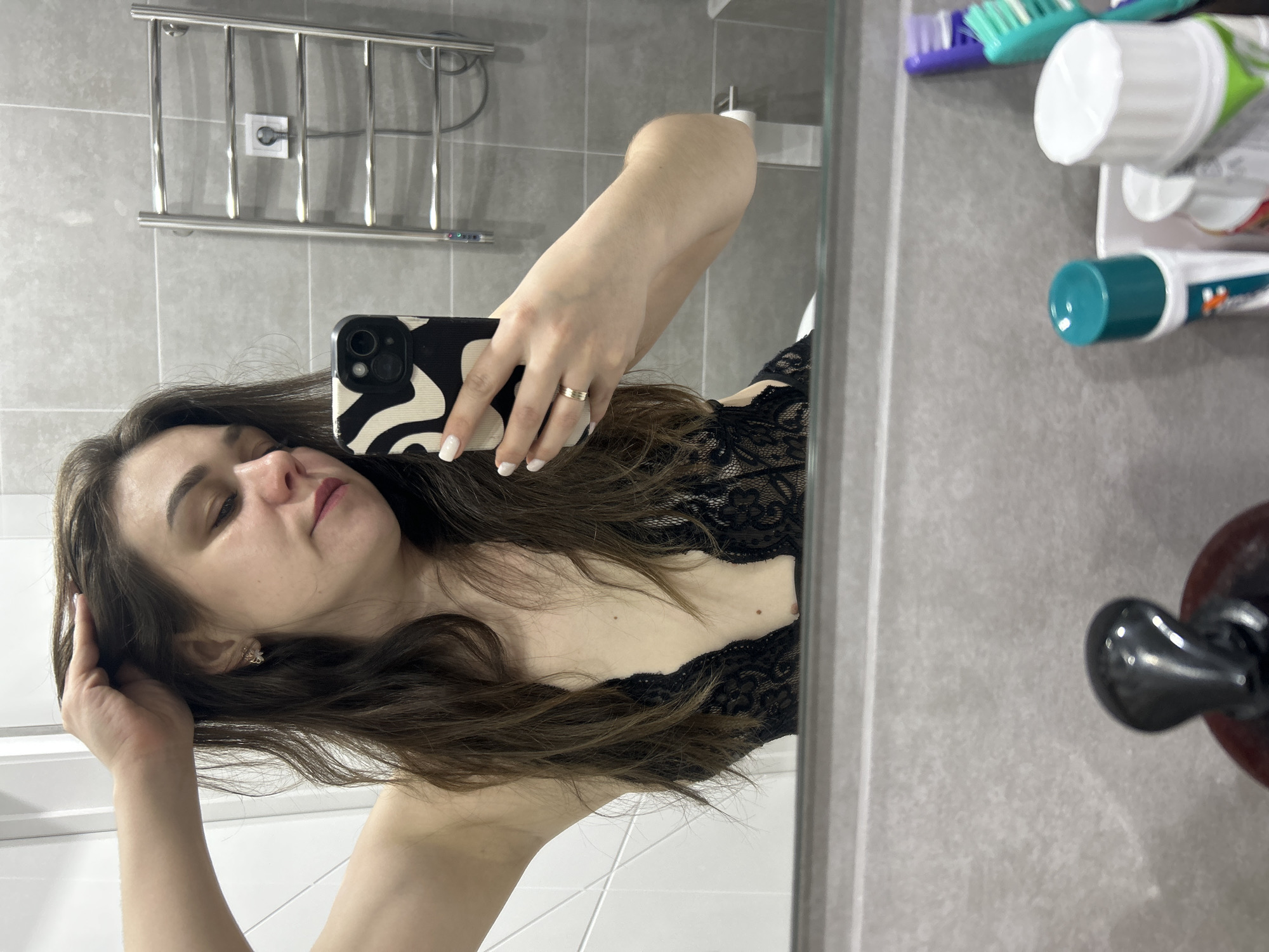 An indoor mirror selfie captures a young woman in a bathroom setting. She has long, dark brown hair, which she touches with her right hand near her head. Her left hand, adorned with white nail polish and a simple band ring on the ring finger, holds a smartphone with a distinctive black and white abstract-patterned case. The phone's dual cameras are visible. She is wearing a black lace top or bra. Her expression is calm, with her eyes slightly downcast.
The background, reflected in the mirror, shows a contemporary bathroom with light gray tiled walls and a white tiled floor. A chrome towel warmer/rack is mounted on the wall behind her, with an electrical outlet visible beneath it. To the right, various toiletries are arranged on a shelf or holder, including toothbrushes (one green, one purple) and several tubes of toothpaste, though no readable text is visible on them. A dark, possibly red or brown, object with a black knob, possibly part of a sink or faucet, is partially visible in the lower right corner of the reflection. The overall lighting suggests artificial indoor light.