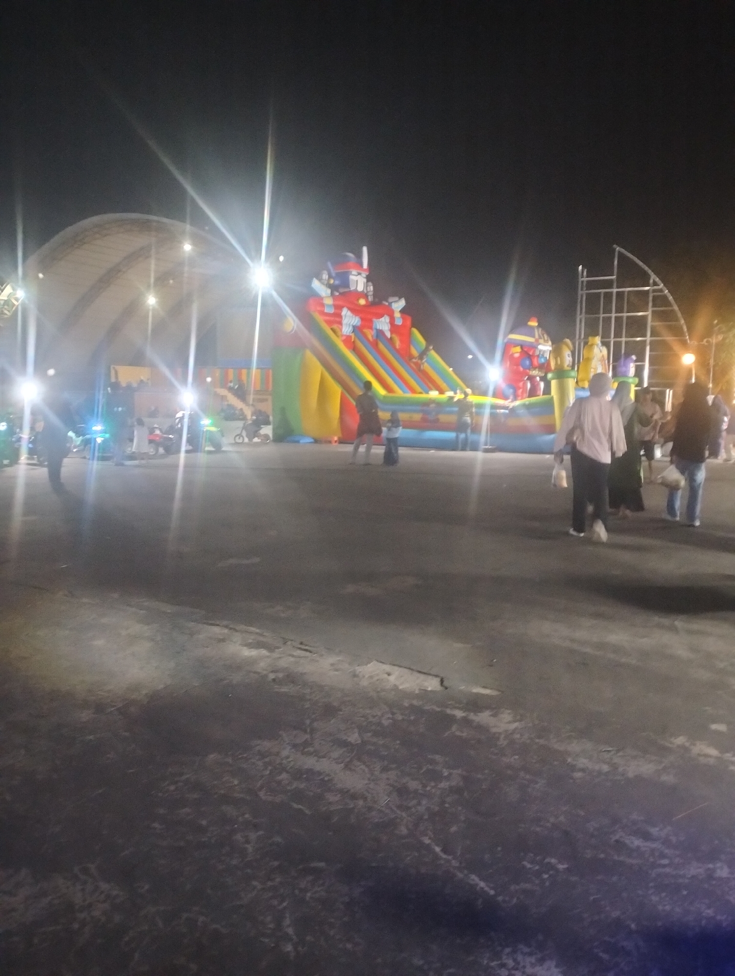 The image depicts an outdoor night scene, possibly a temporary fair or event, in Dumai City, Indonesia. In the background, a large inflatable slide with a "Transformers" theme is visible, featuring characters like Optimus Prime. Several people are present, some walking on a paved surface, while others appear to be watching or participating in activities. In the foreground, there are several small motorized vehicles, likely for children's entertainment, with bright lights. The overall lighting is dim, with harsh artificial lights creating lens flares. A large, dome-like structure with a white roof is partially visible on the left side of the image. The ground is a dark, textured pavement.
