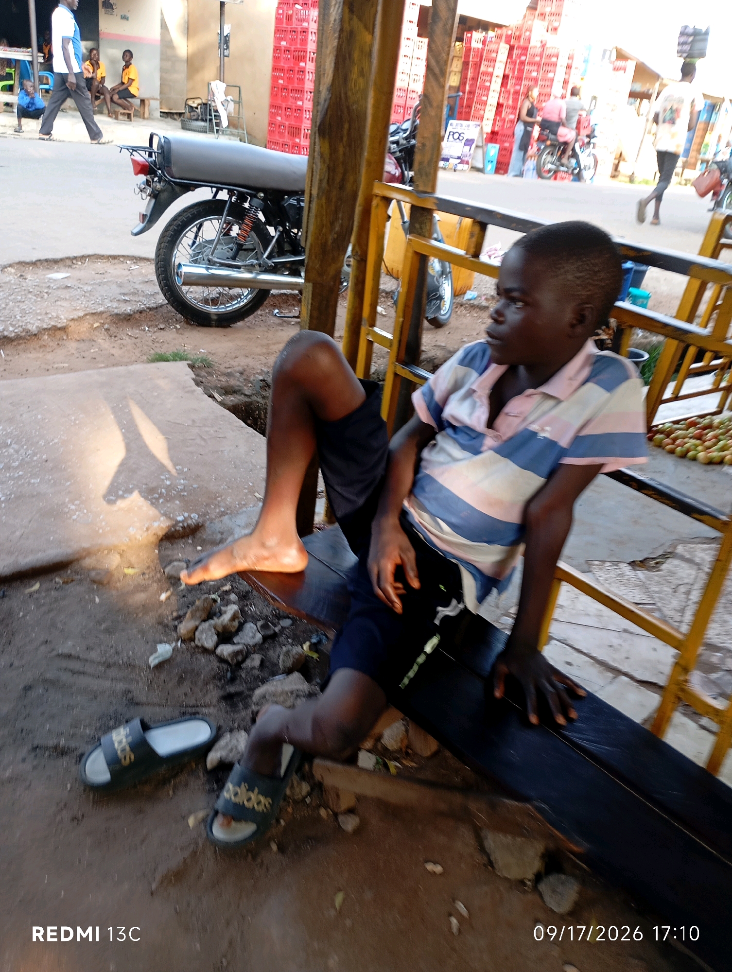 A young boy with dark skin and short hair sits on a dark wooden bench, looking to his right. He is wearing a striped polo shirt with pink and blue horizontal bands and dark blue shorts. His left leg is extended, with his bare left foot resting on the ground. A pair of black flip-flops with gold lettering are on the ground near his feet. The scene appears to be outdoors in an urban or semi-urban environment, possibly a market or street. Behind the boy, a motorcycle is parked, and stacks of red crates are visible in the background, along with other people and buildings. The ground is a mix of dirt and concrete, with some scattered rocks. The lighting suggests it is daytime, with sunlight casting shadows. The timestamp "09/17/2026 17:10" is visible in the lower right corner. The location is identified as Shendam, Nigeria.